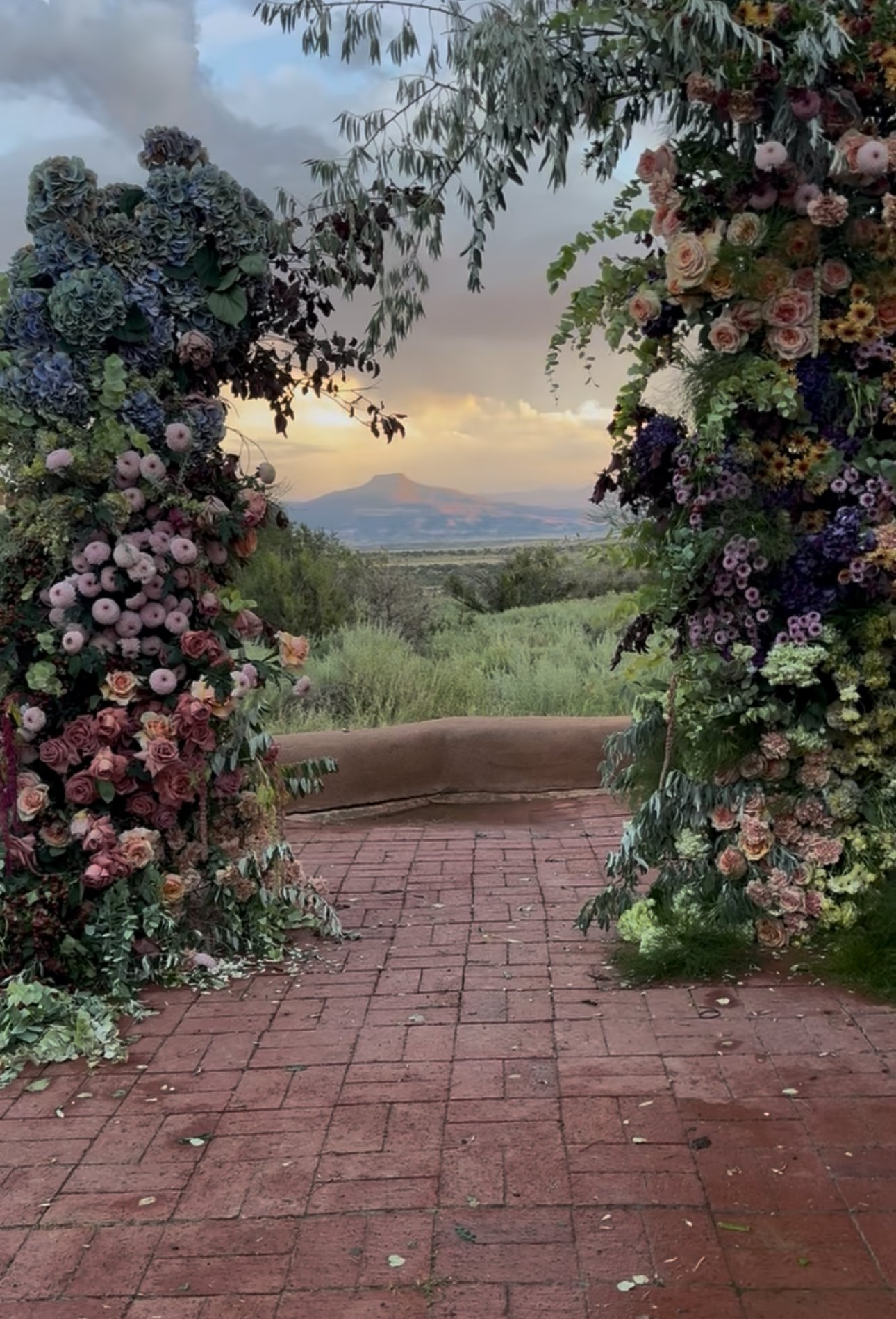 A floral archway with pink, purple, and blue flowers, framing a view of a distant mountain at sunset over a lush green landscape.