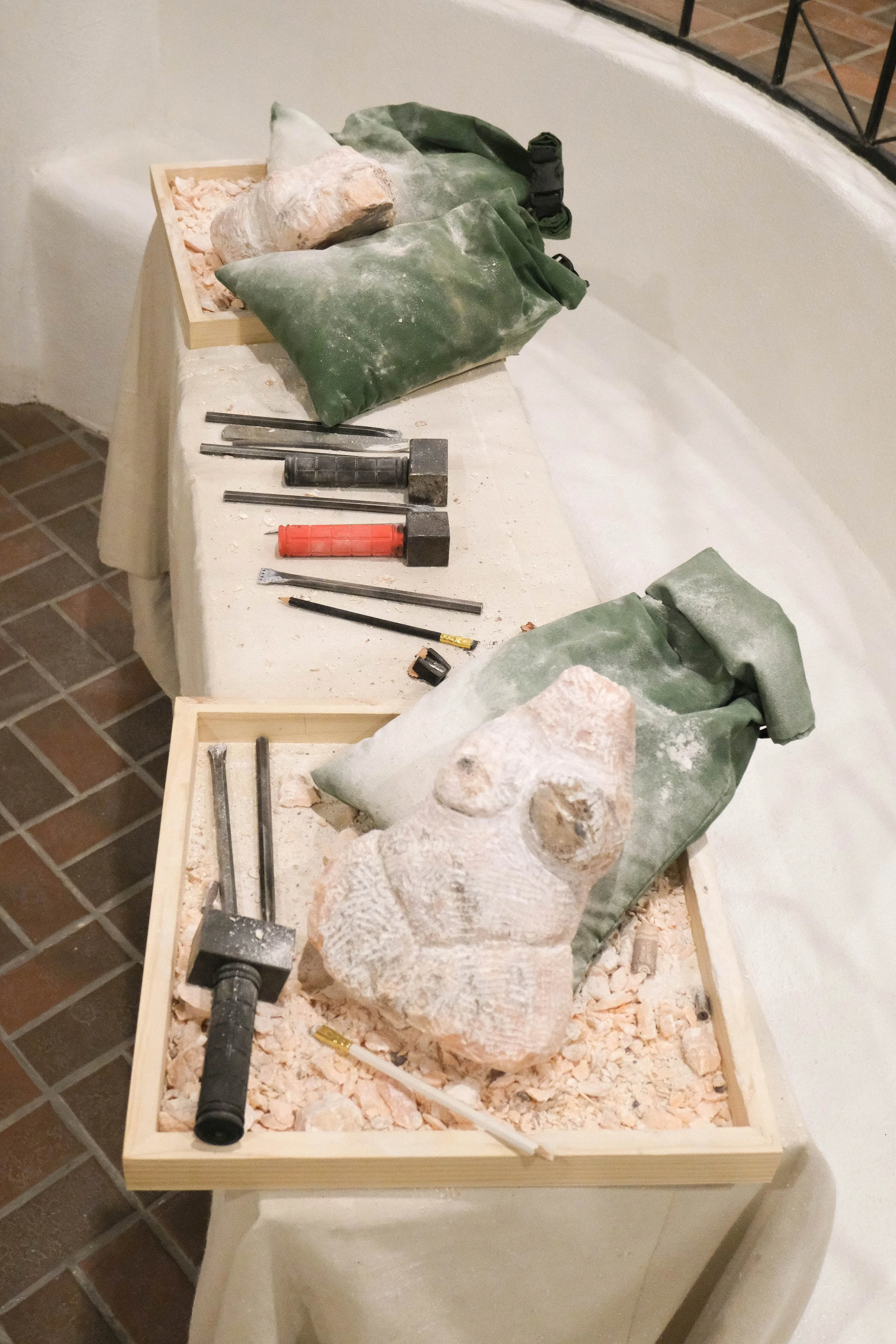 Display of sculpting tools, limestone, and green bags on tables, with a partially carved limestone sculpture in a museum or gallery setting.