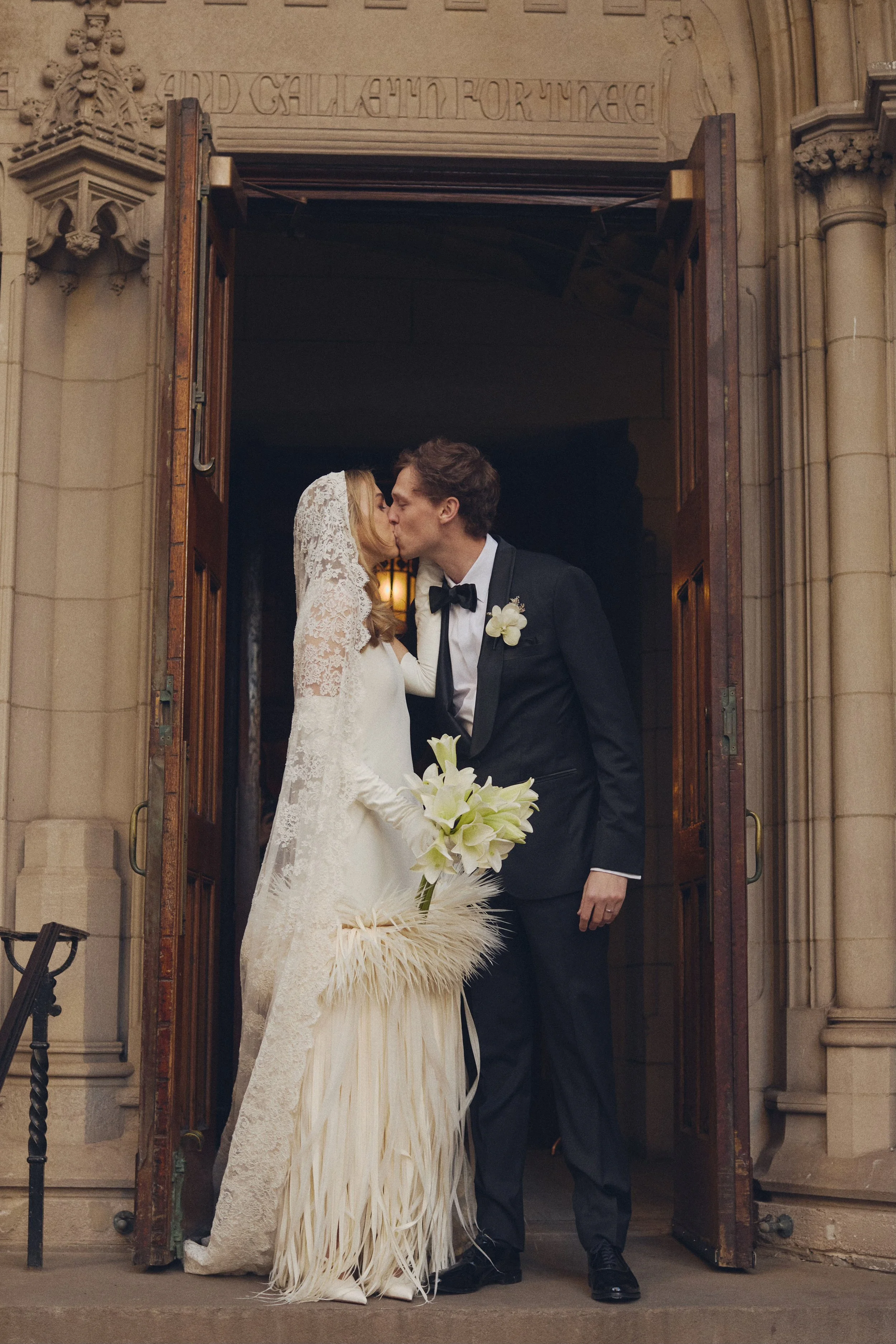 Bride and groom kissing at the church entrance, with the bride holding a bouquet of white flowers, wearing a lace wedding dress and veil, and the groom in a tuxedo with a black bow tie and white boutonniere.