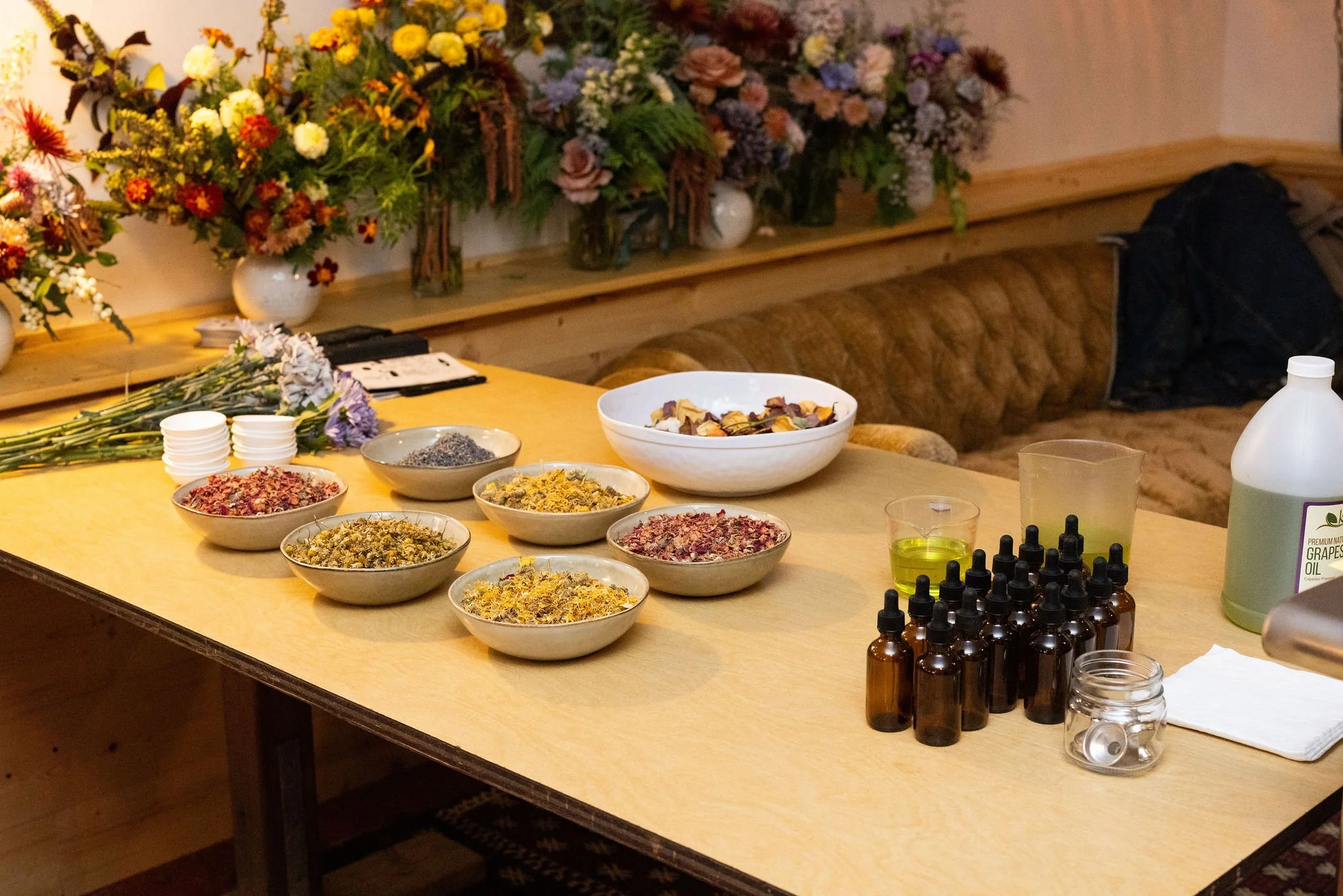 A table with various bowls of dried herbs and flowers, small bottles with droppers, a container of grape seed oil, a glass of yellow oil, and a jar of liquid, with a backdrop of colorful flower arrangements.