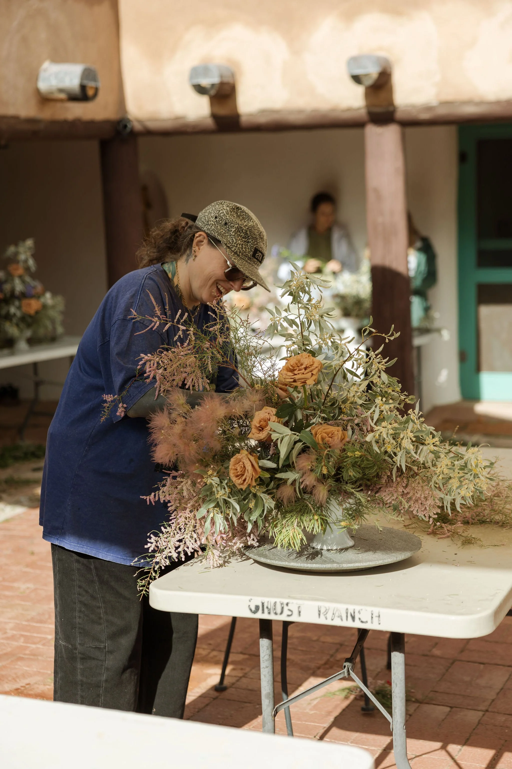 A woman wearing sunglasses, a leopard-print hat, and a blue jacket arranging a floral display at an outdoor event, with a table labeled 'GHOST RANCH' in front of her.