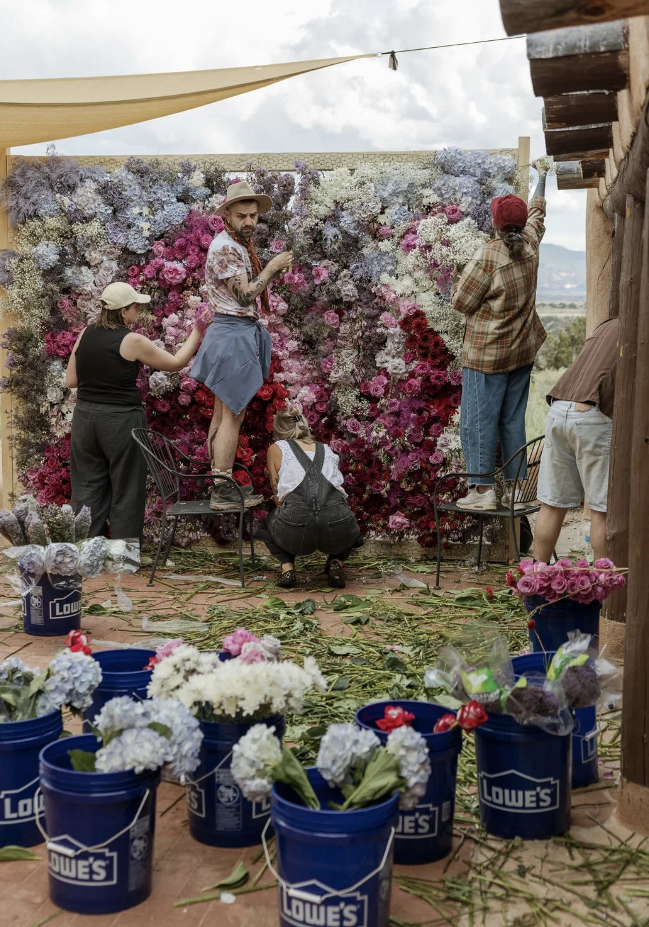 People assembling a large floral backdrop with pink, white, and purple flowers, with blue Lowe's buckets and fallen flowers on the ground.