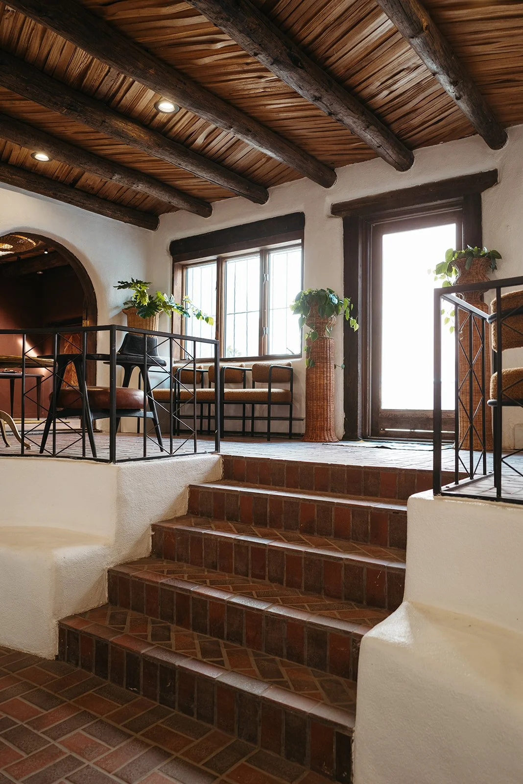 Interior of a rustic apartment with brown tiled stairs, white textured walls, wooden ceiling beams, and large windows with potted plants.