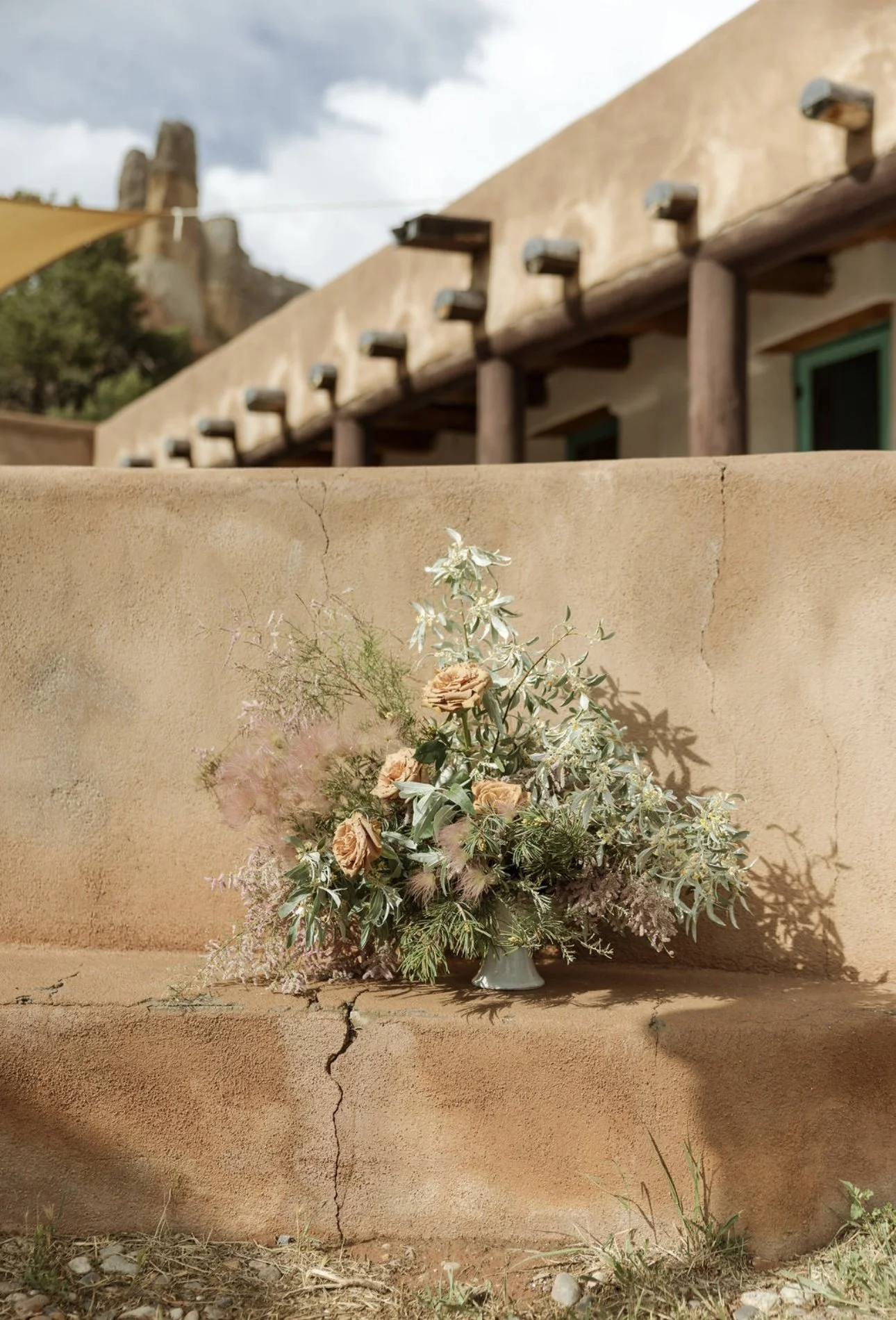 A flower arrangement in a small white vase placed on a cracked adobe wall, with a rustic building and a cloudy sky in the background.