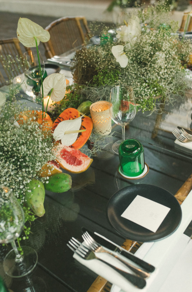 A decorated dining table featuring floral arrangements with white flowers and greenery, cut citrus fruits including grapefruit and orange, a green candle, an empty wine glass, and cutlery set on a white napkin.