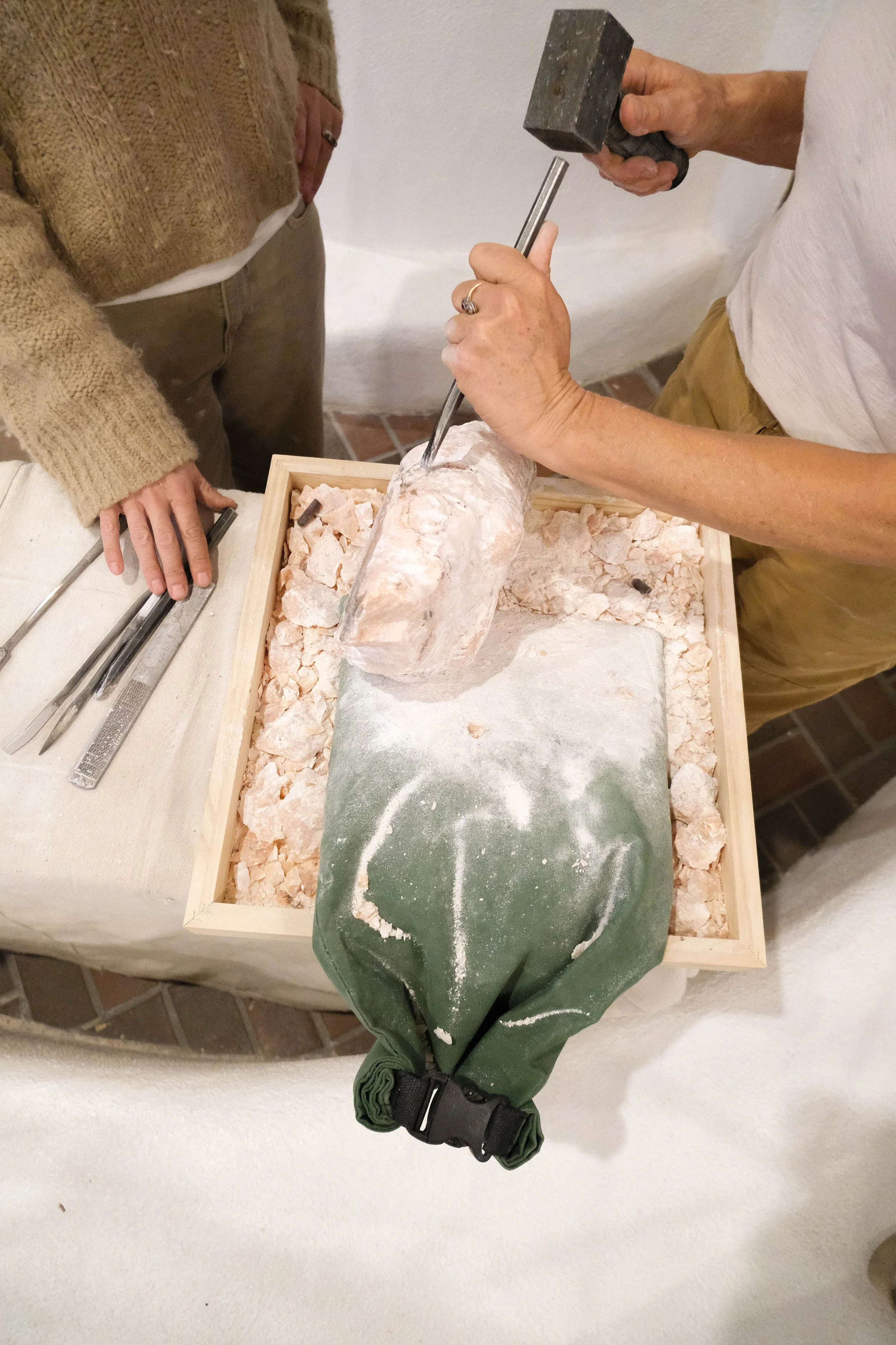Person holding a hammer and chisel, working on a large salt block or mineral specimen in a wooden box, with another person observing nearby.
