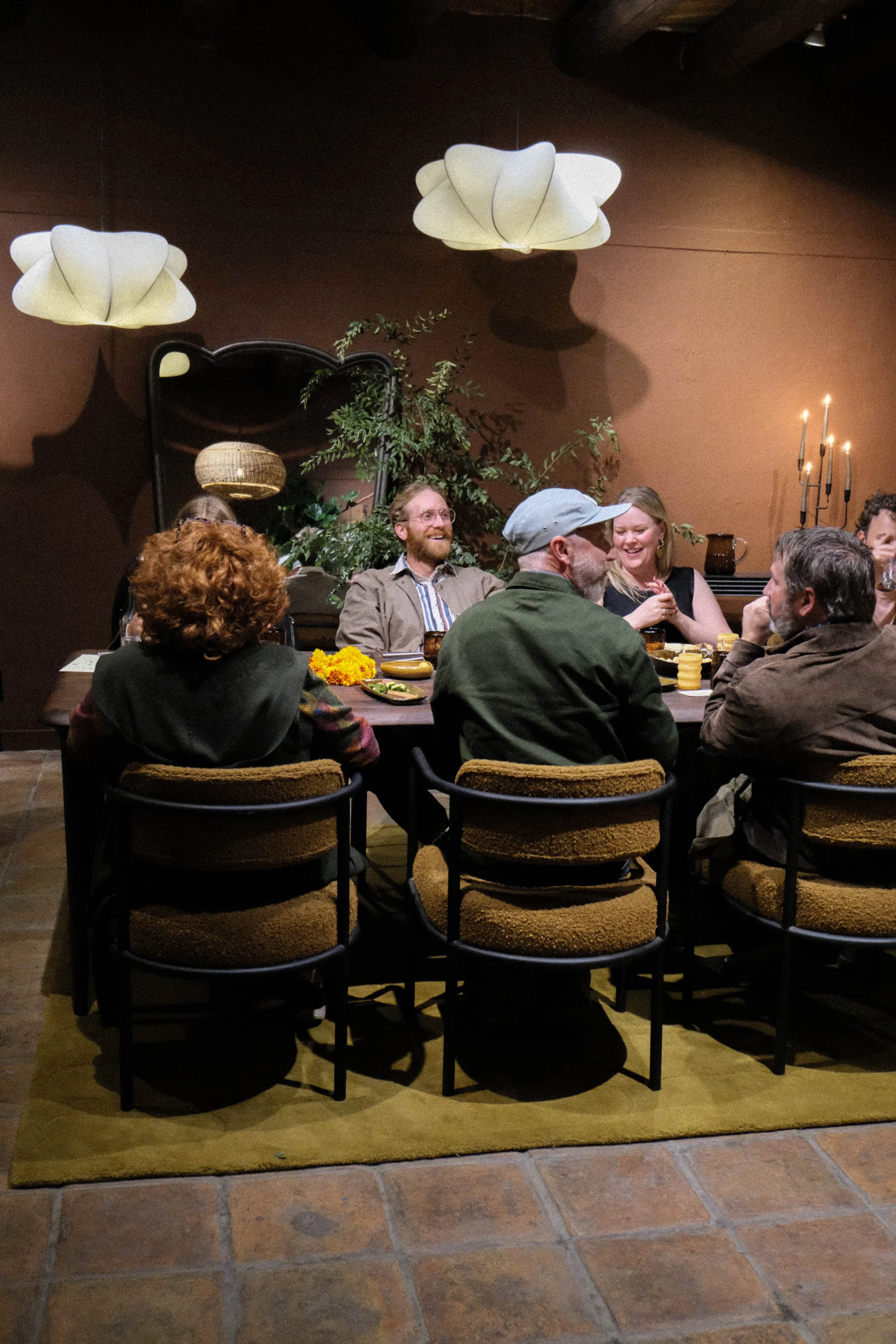 People sitting at a dining table in a cozy restaurant, engaging in conversation and laughter, with warm lighting and decorative plants in the background.