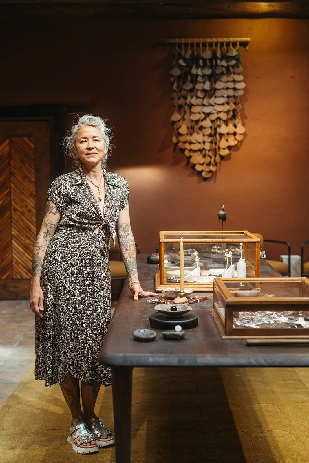 A woman with tattoos on her arms standing next to a wooden table with display cases, candles, and small objects inside, in a room with brown walls and a decorative art piece hanging on the wall behind her.