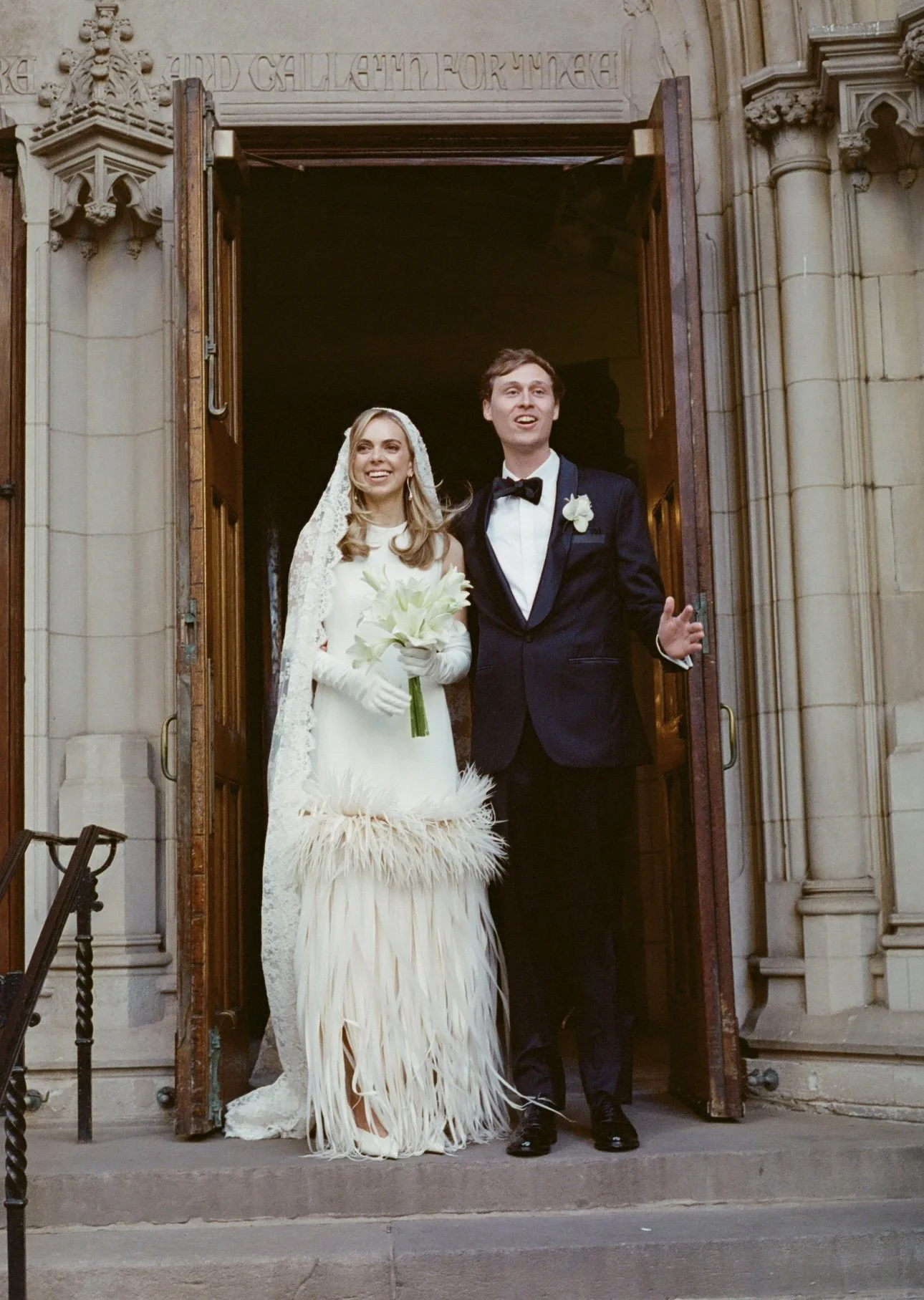 A bride and groom standing at church entrance, smiling after their wedding, bride holding a bouquet of white flowers, groom in tuxedo with a bow tie and boutonniere.
