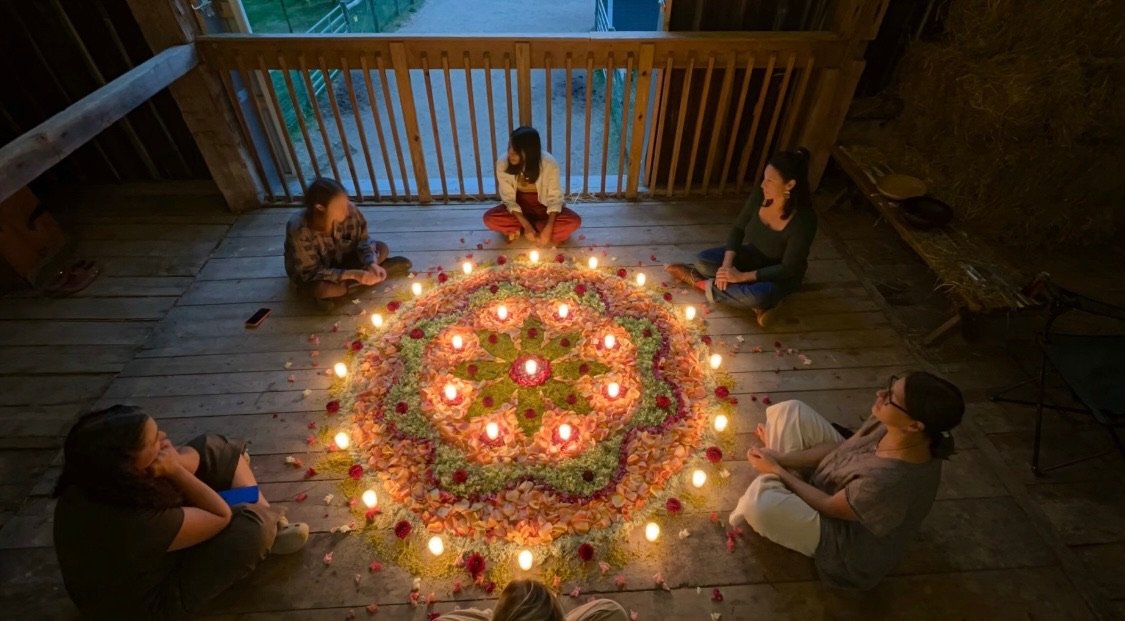 Six women sitting in a circle around a large flower-shaped floral arrangement with lit candles and rose petals on a wooden deck.