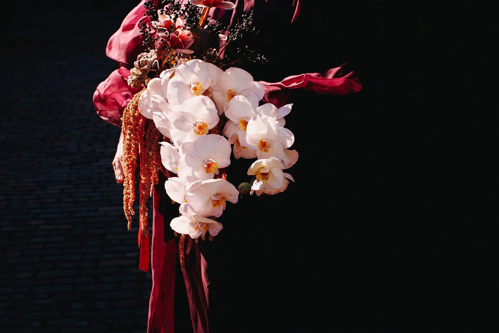 A bouquet of white orchids with pink and red flowers, wrapped with red ribbons, against a dark background.