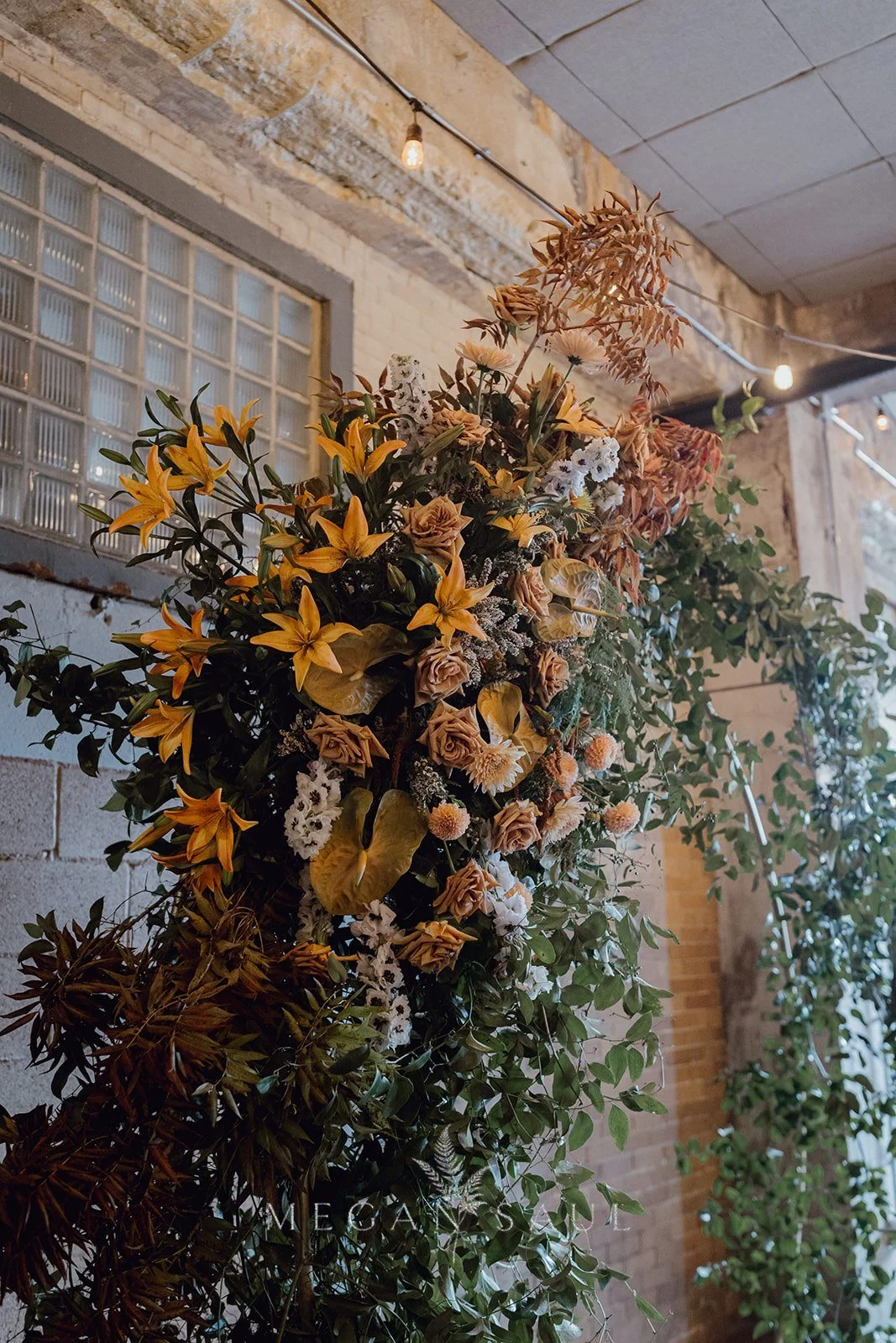 A large hanging floral arrangement with yellow lilies, roses, and various dried flowers and green foliage, hanging inside a rustic brick and wood interior with string lights.