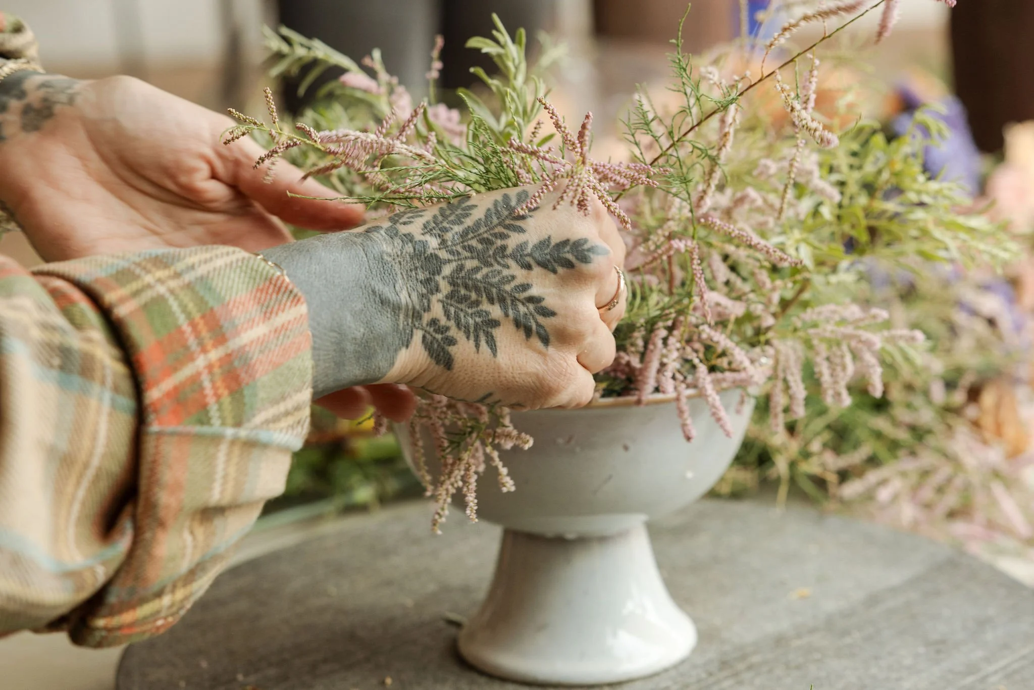 Person with tattooed hand arranging pink and green floral arrangement in a white pedestal bowl.