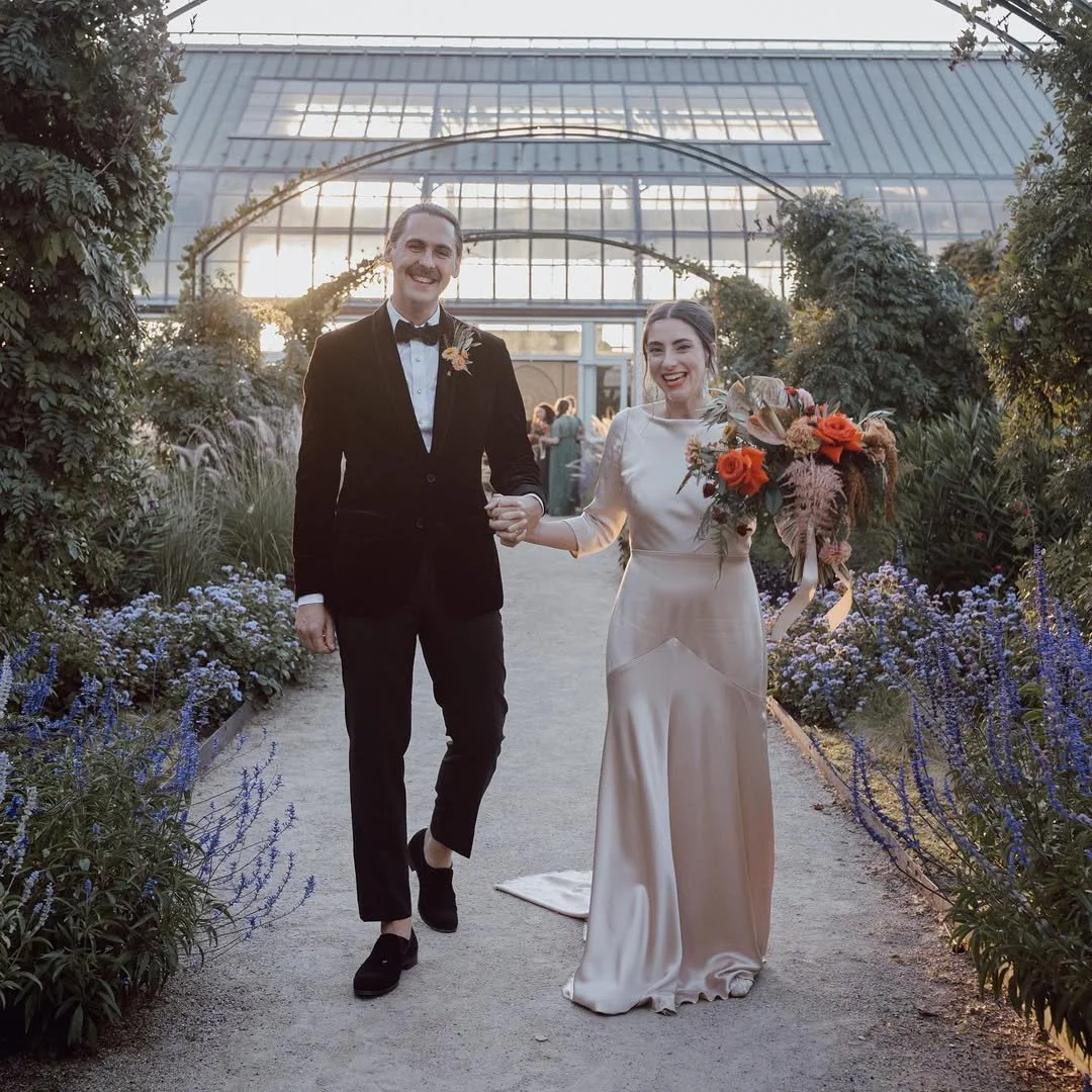 A happy couple, dressed in wedding attire, holding hands and smiling while walking down a garden path with purple flowers on either side, inside a greenhouse with sunlight streaming through the glass roof.
