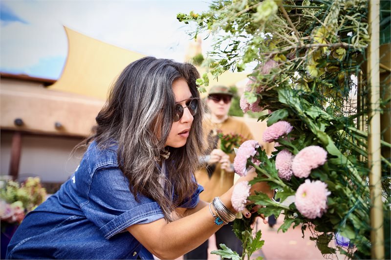 A woman with long dark hair and sunglasses is adjusting or inspecting pink and white flowers on a garden trellis, with another person wearing sunglasses and a hat in the background.