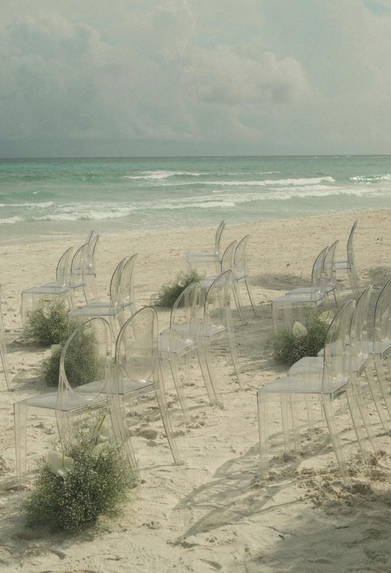 Empty transparent chairs arranged on a sandy beach with small green plants, facing the ocean. Overcast sky with clouds.