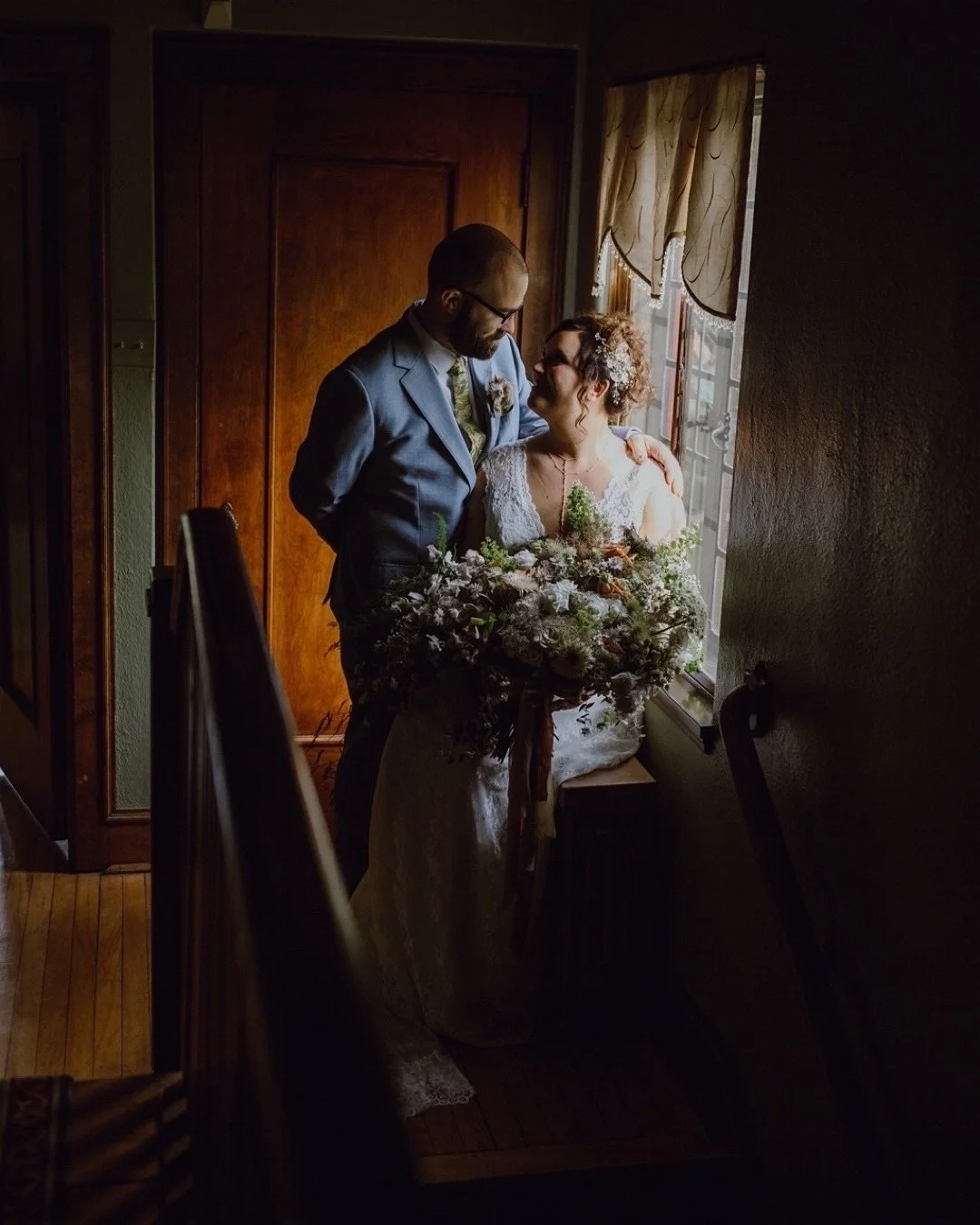 A bride and groom sharing an intimate moment near a window, with the bride holding a bouquet and both dressed in wedding attire, in a dimly lit room.