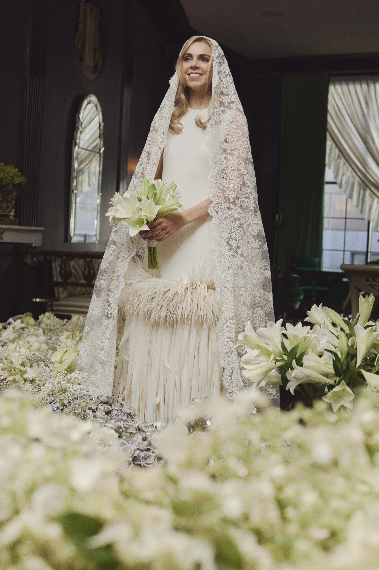 A bride in a white wedding dress holding a bouquet of white calla lilies, standing surrounded by white flowers and greenery, inside a decorated venue.