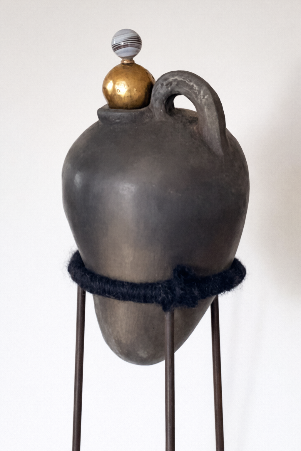 A ceramic vessel with a rounded body, a small handle, and a narrow opening, embroidered with a black band and topped with three decorative balls of different sizes and materials, supported by metal legs.