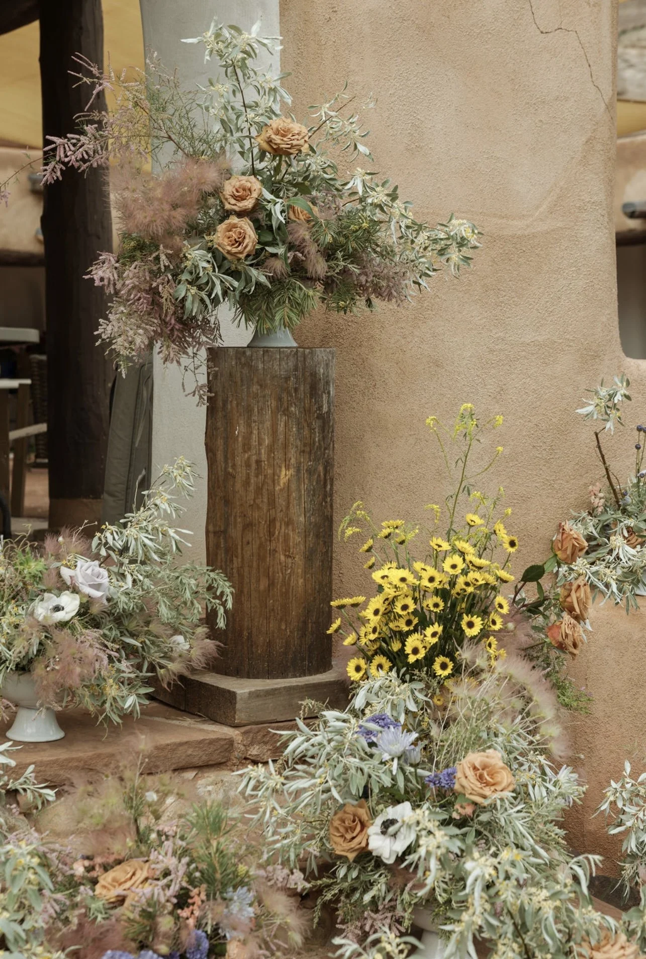 A decorative floral arrangement with peach roses, white flowers, and pinkish-lavender plants in a white vase, placed on a tall wooden pedestal with yellow flowers nearby, against a beige stucco wall.