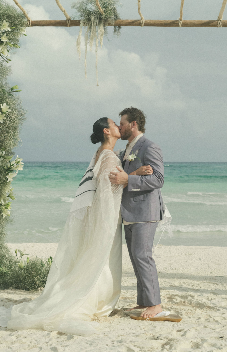 A bride and groom sharing a kiss at their beach wedding ceremony, standing on sand with the ocean in the background, under a wooden arch decorated with greenery and white flowers.