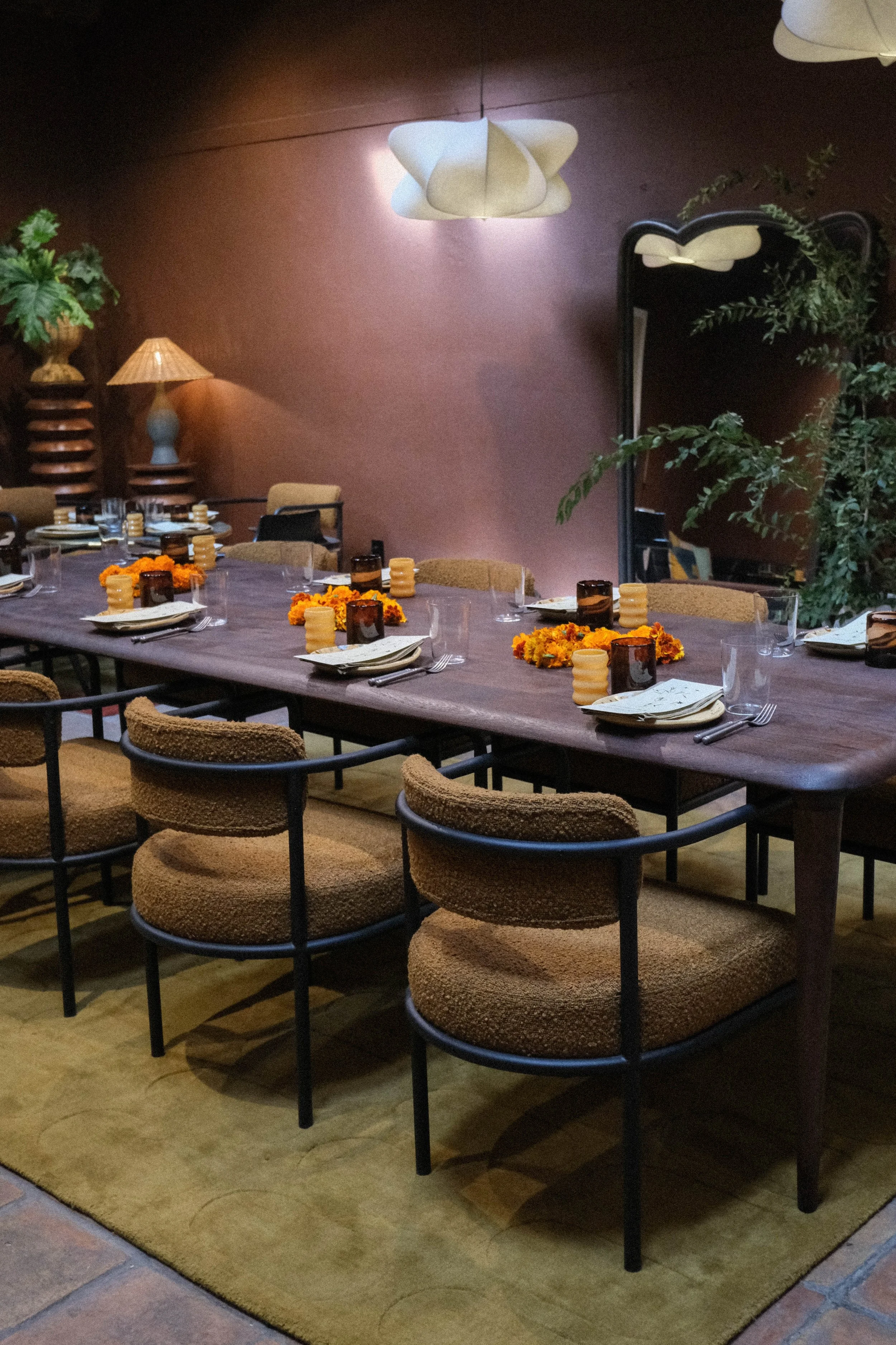 A long dining table set for a meal with plates, glasses, silverware, napkins, and centerpieces made of orange marigold flowers. The table is surrounded by brown upholstered chairs with black metal frames. The room has warm lighting, a large mirror, p