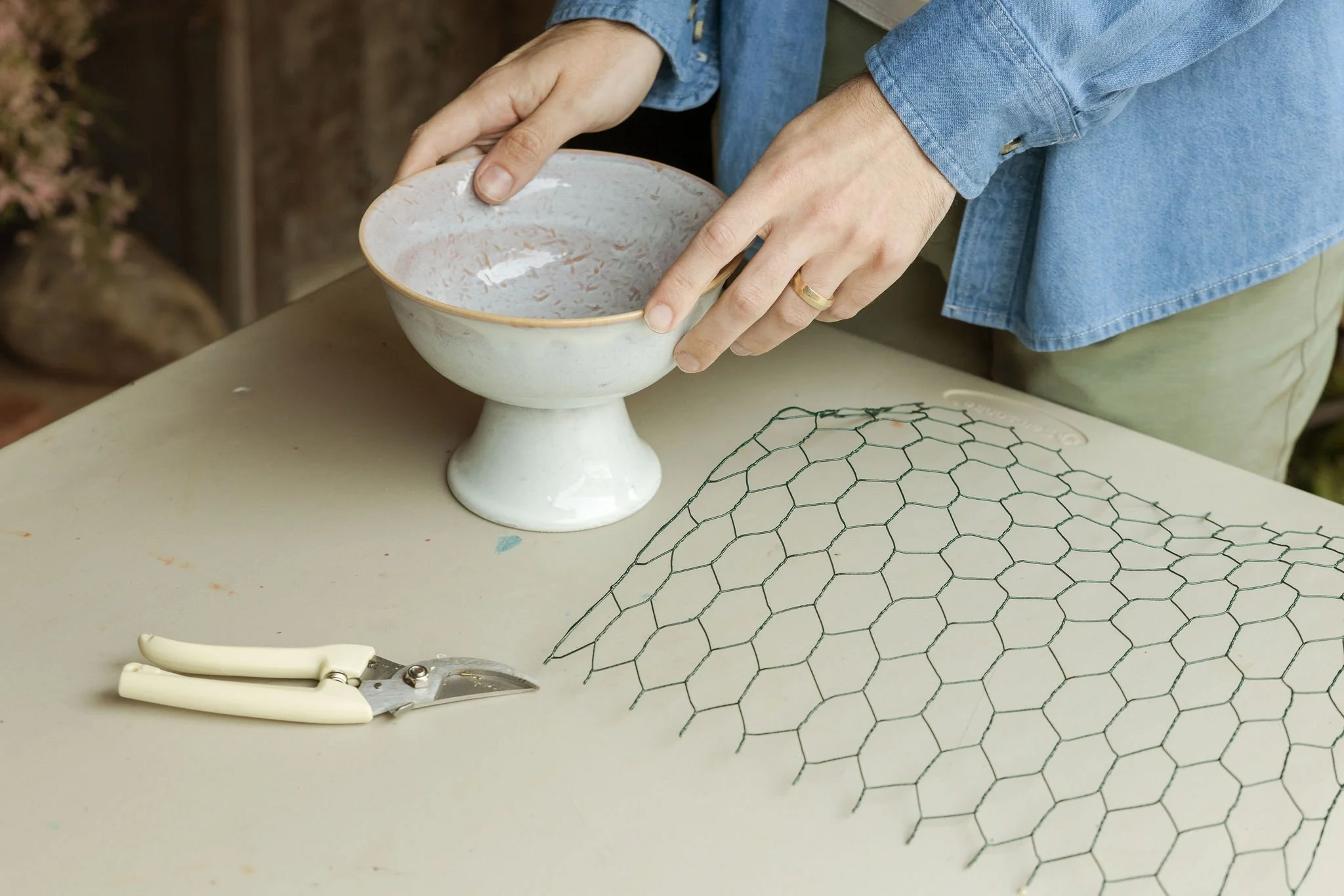 Person working on garden art, shaping a wire frame with hexagonal patterns, with a ceramic bowl and gardening shears on the table.