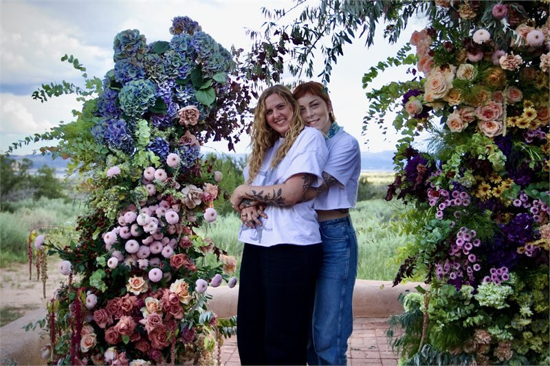 Two women hugging and smiling in front of a floral arch outdoors with green field and cloudy sky in the background.