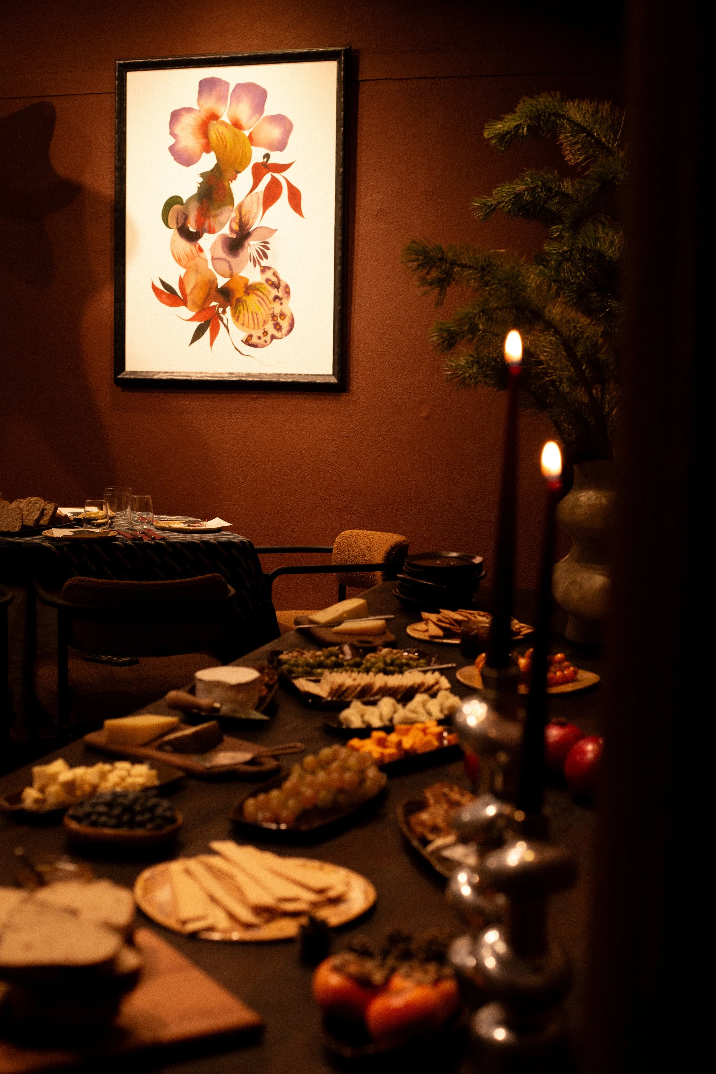A dimly lit dining room with a table of assorted cheeses, crackers, and fruits, a wall decor featuring framed floral art, and a vase with pine branches.