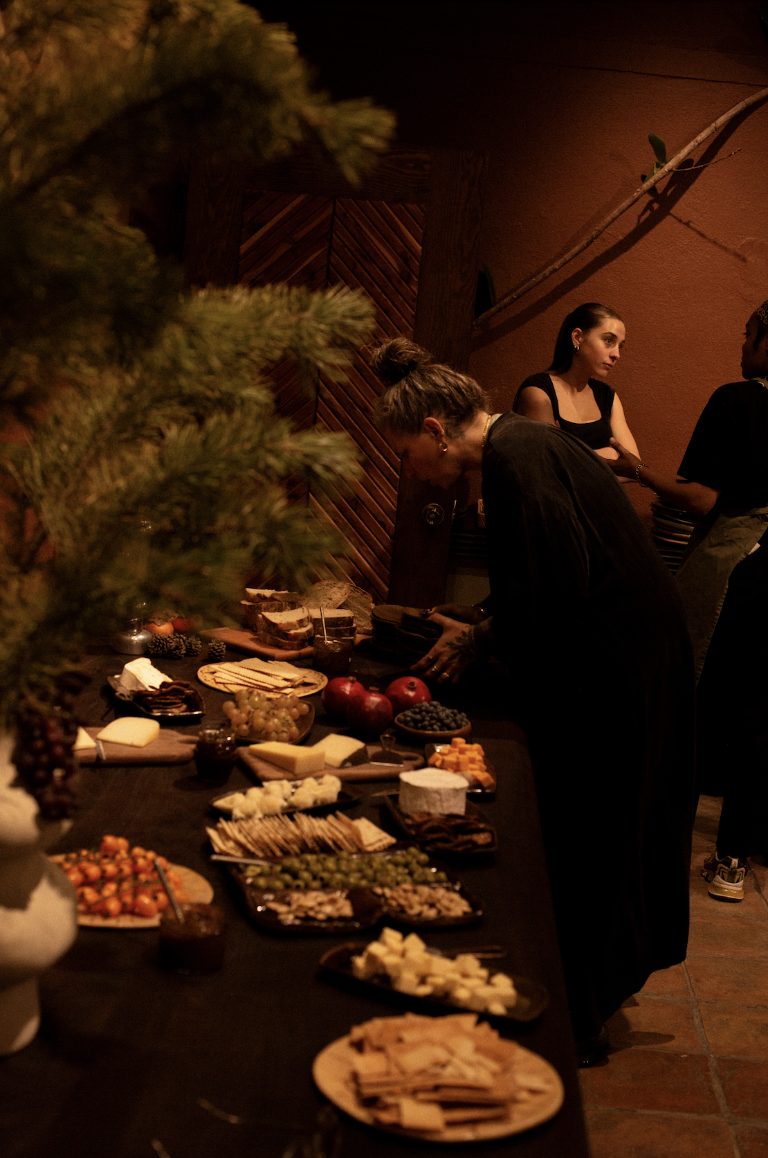 A buffet table with various cheeses, fruits, meats, and crackers, with people serving themselves in a dimly lit room.