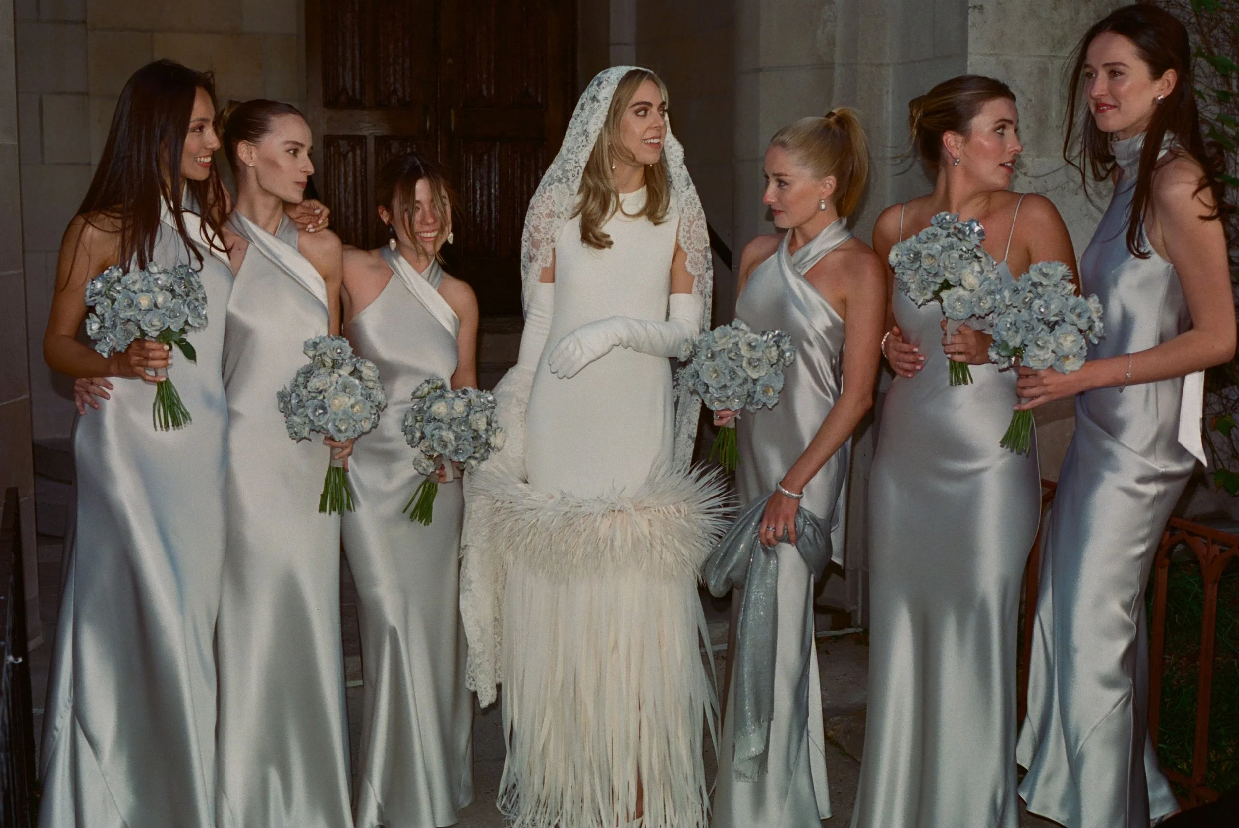 Bride in a white wedding dress surrounded by bridesmaids in silver gowns holding bouquets