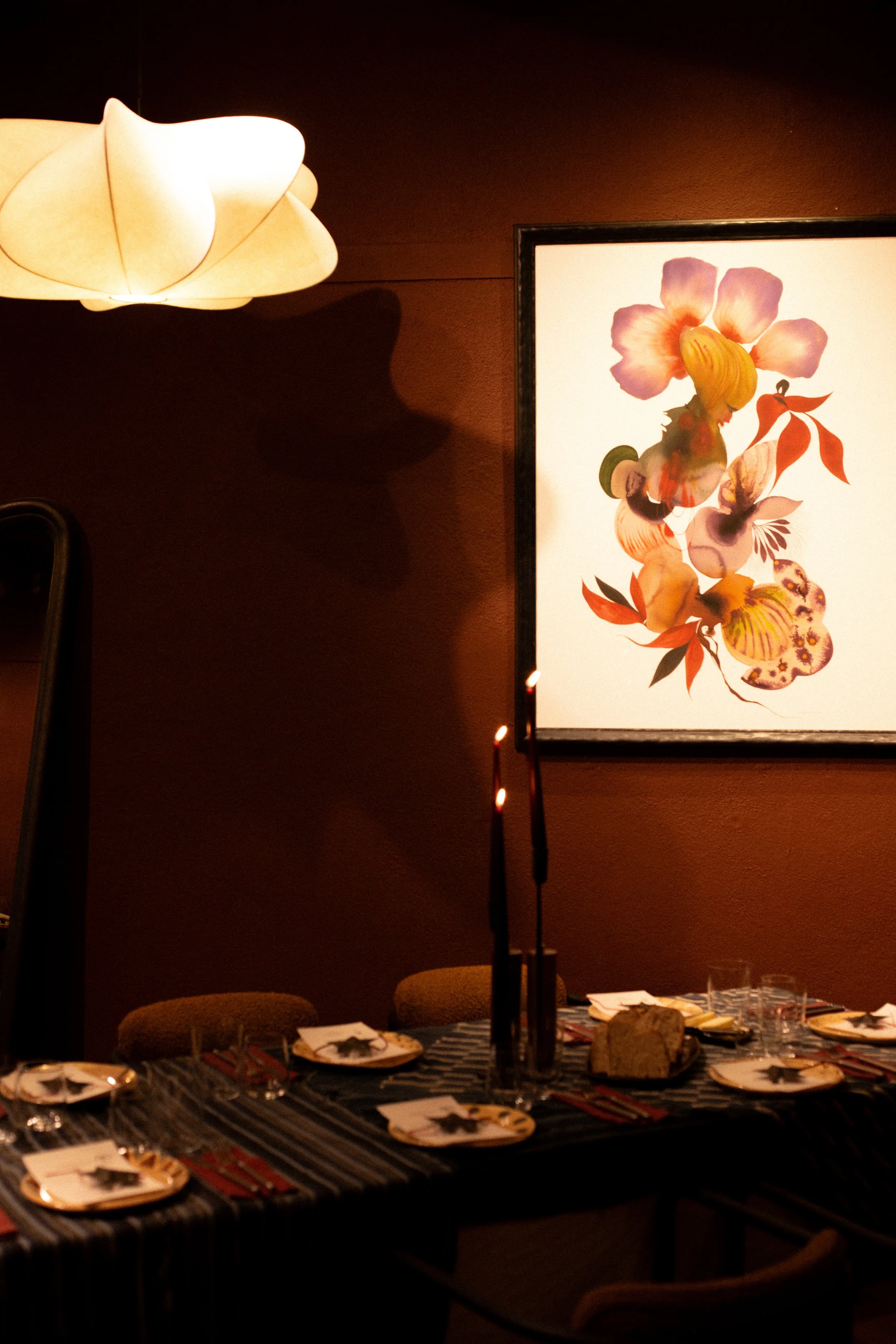 A dining table set for multiple guests with plates, glasses, and cutlery, decorated with a tall candle holder with lit candles and a large bread loaf, in a dimly lit room with a floral artwork on the wall and a modern ceiling lamp casting shadows.