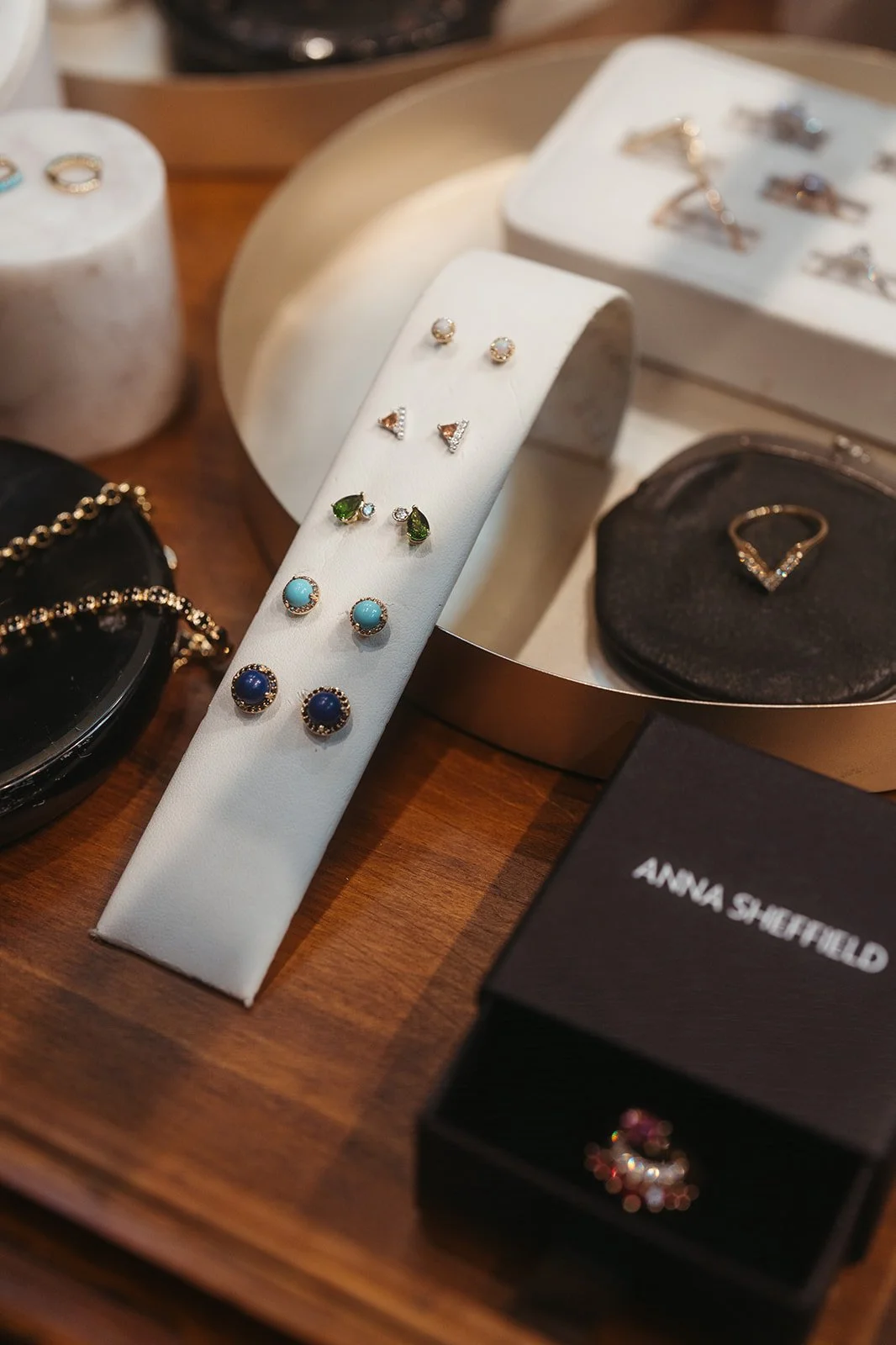 Display of various earrings, including stud, triangular, teardrop, and gemstone designs, arranged on a white stand with a jewelry box and necklace in the background.