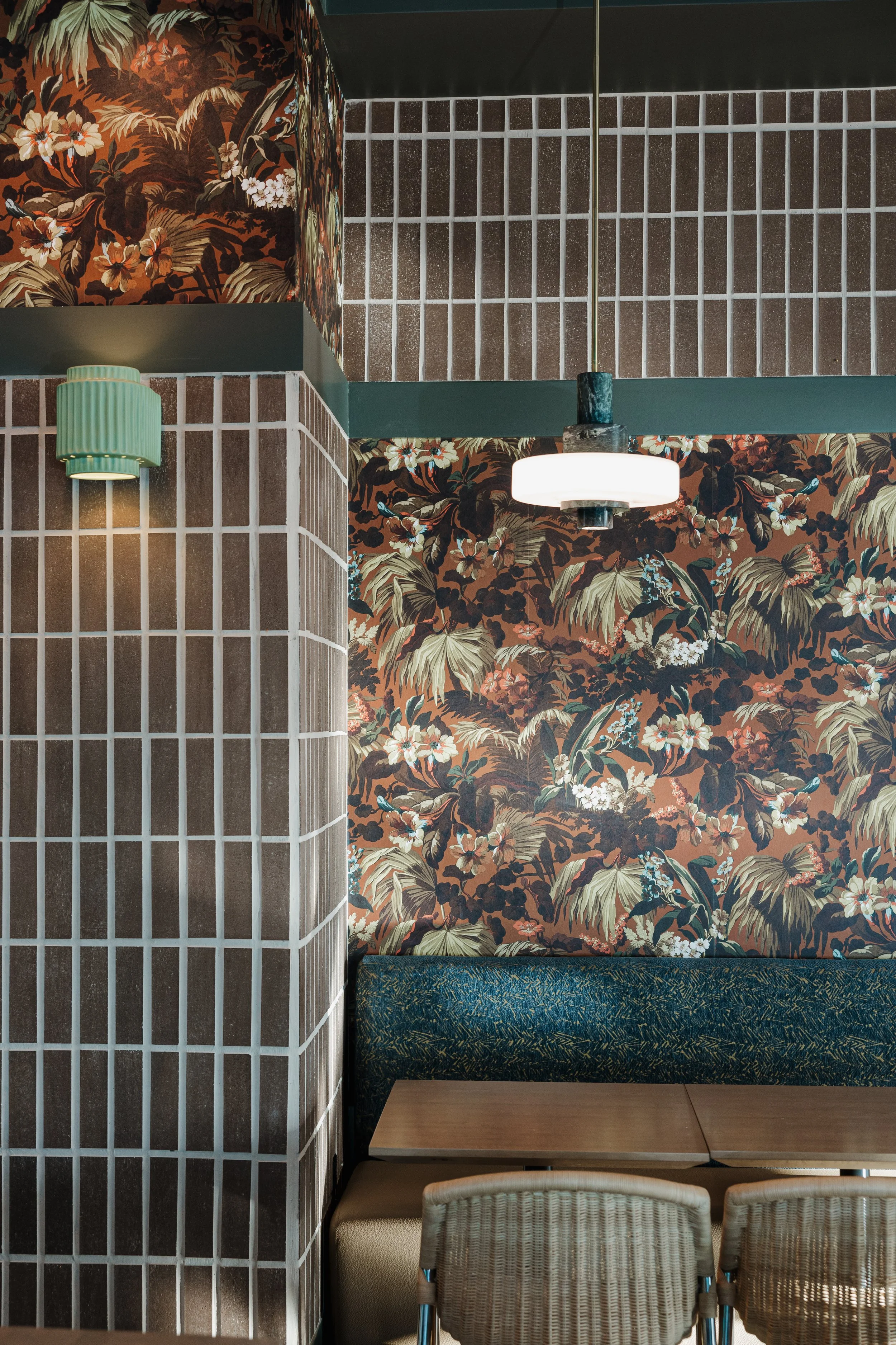 Interior of a restaurant or cafe with floral wallpaper, a dark-blue cushioned bench, a light wood table, rattan chairs, a green wall sconce, and a ceiling light fixture.