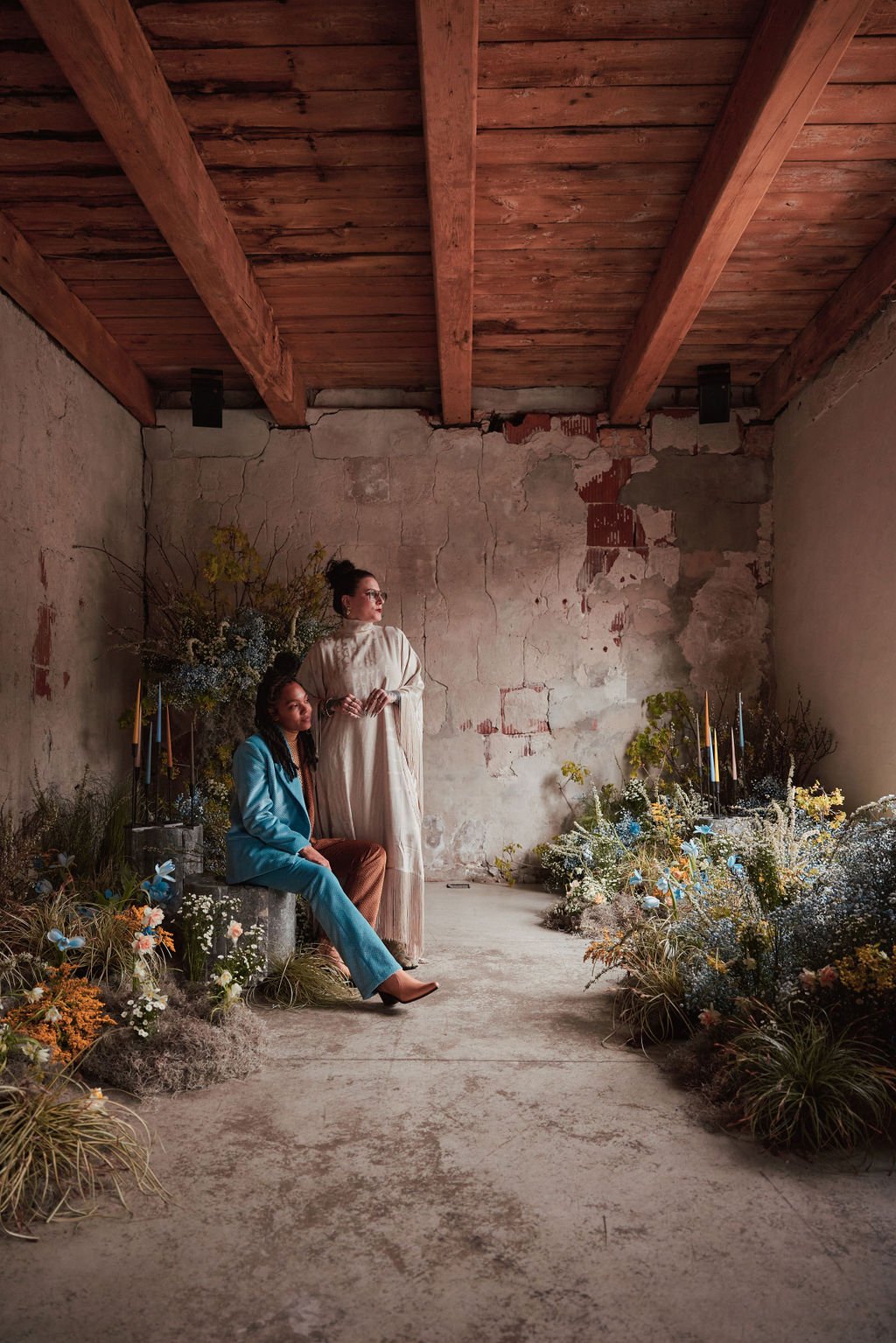 Two women in vintage clothing in a rustic room with a weathered wall and blooming flowers, one sitting on a stone, the other standing beside her.
