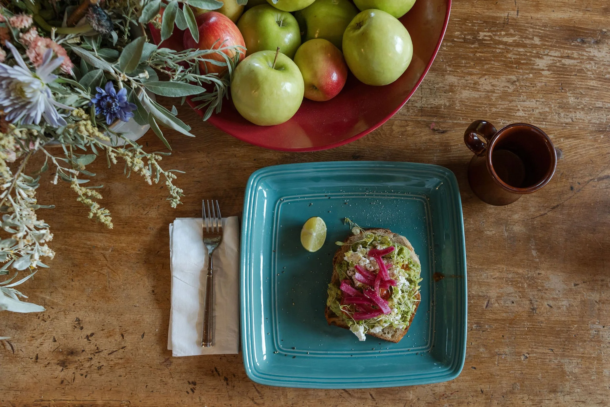 A table with a slice of avocado toast topped with microgreens and pickled red onions on a blue plate, a wedge of lime, a fork on a napkin, a large bowl of green apples and red apples, and a brown mug on a wooden table.