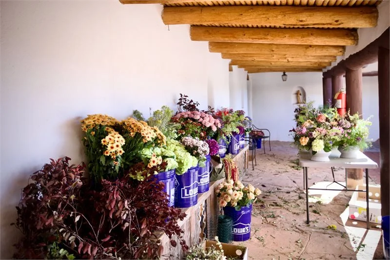 Flower arrangements placed on tables and buckets along a corridor with wooden ceiling beams.