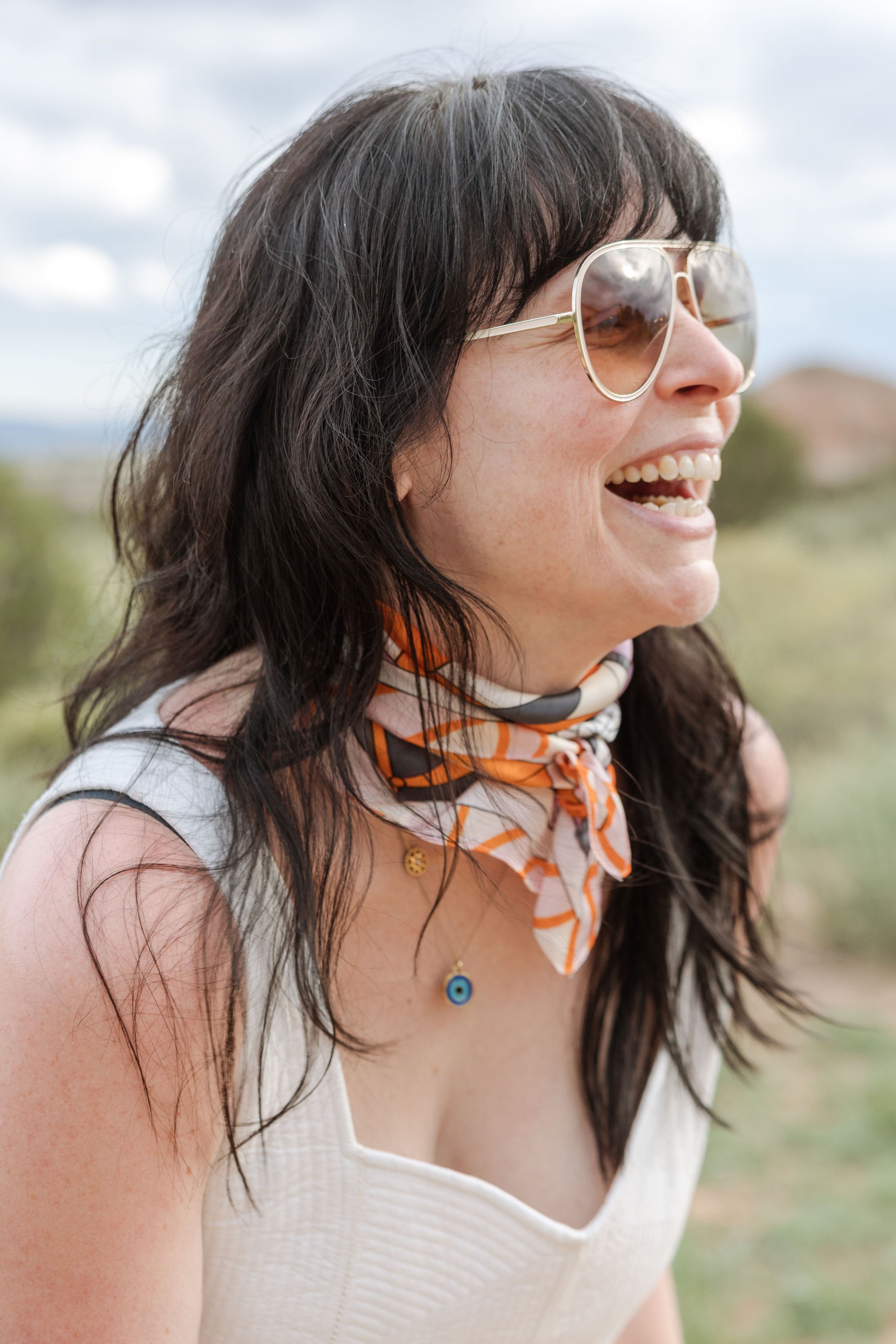 Smiling woman wearing sunglasses, a white sleeveless top, an orange and white striped scarf, and a necklace with a blue evil eye pendant, outdoors with a cloudy sky.