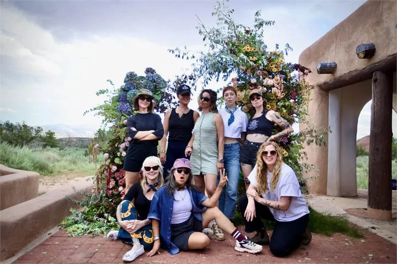 A group of nine women posing outdoors in front of a floral arrangement, with a structure resembling a southwestern style building on the right.