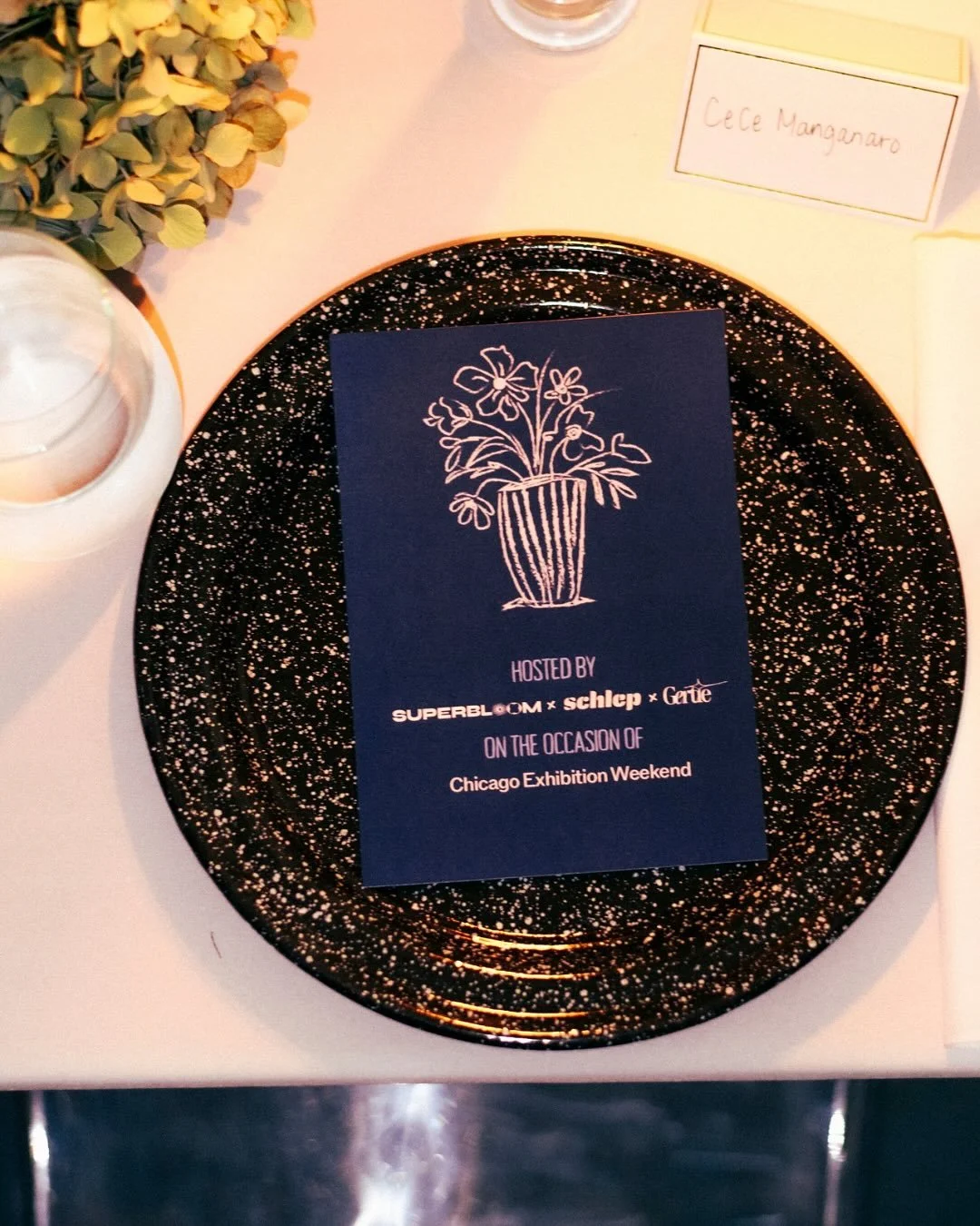 Event invitation card on a black speckled plate. The card features a line drawing of a potted plant with flowers and states it is hosted by Super Bloom, Schlepp, and Gefie during the Chicago Festival Weekend.