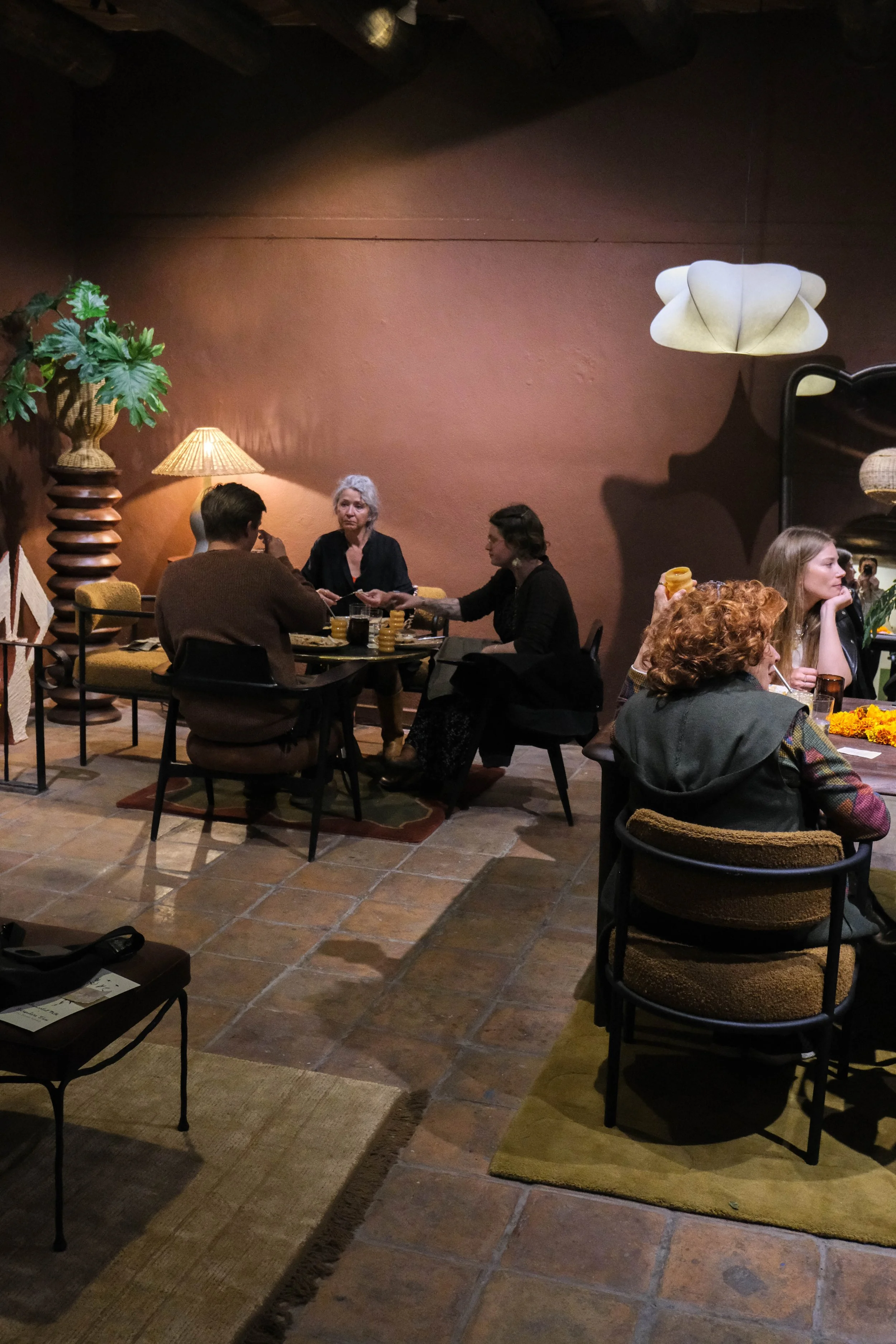 People sitting at tables inside a cozy restaurant or cafe with warm lighting, terracotta tiled floor, and decorative plants and lamps on the wall.
