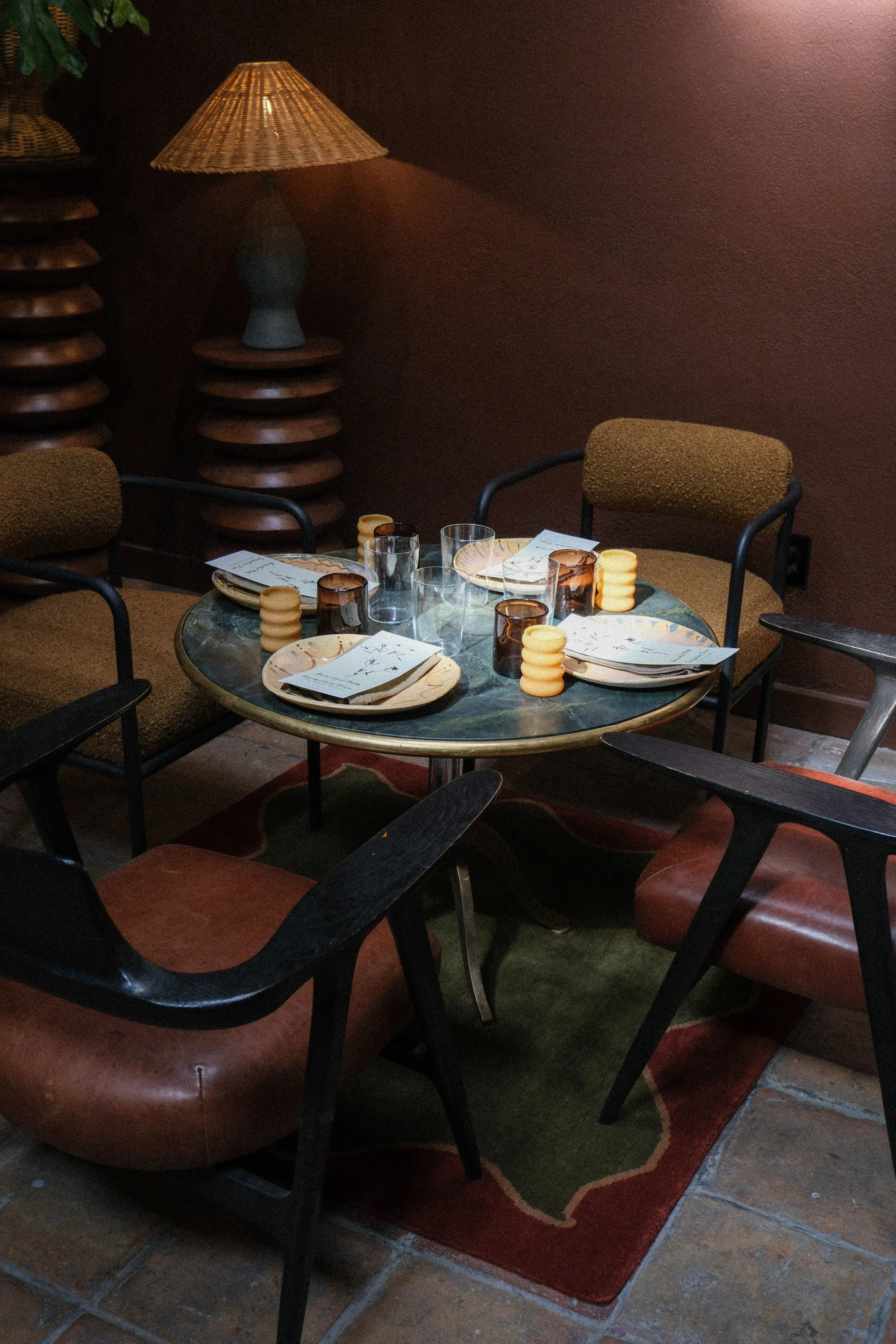 A round dining table set with plates, glasses, candles, and napkins, surrounded by chairs in a dimly lit room with a patterned rug and a brown wall.