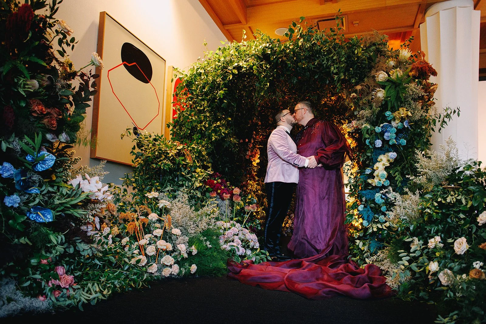 Two men dressed in formal attire, one in a lavender suit and the other in a dark red gown, sharing a kiss while holding hands amidst a backdrop of lush greenery and flowers, in front of abstract artwork.