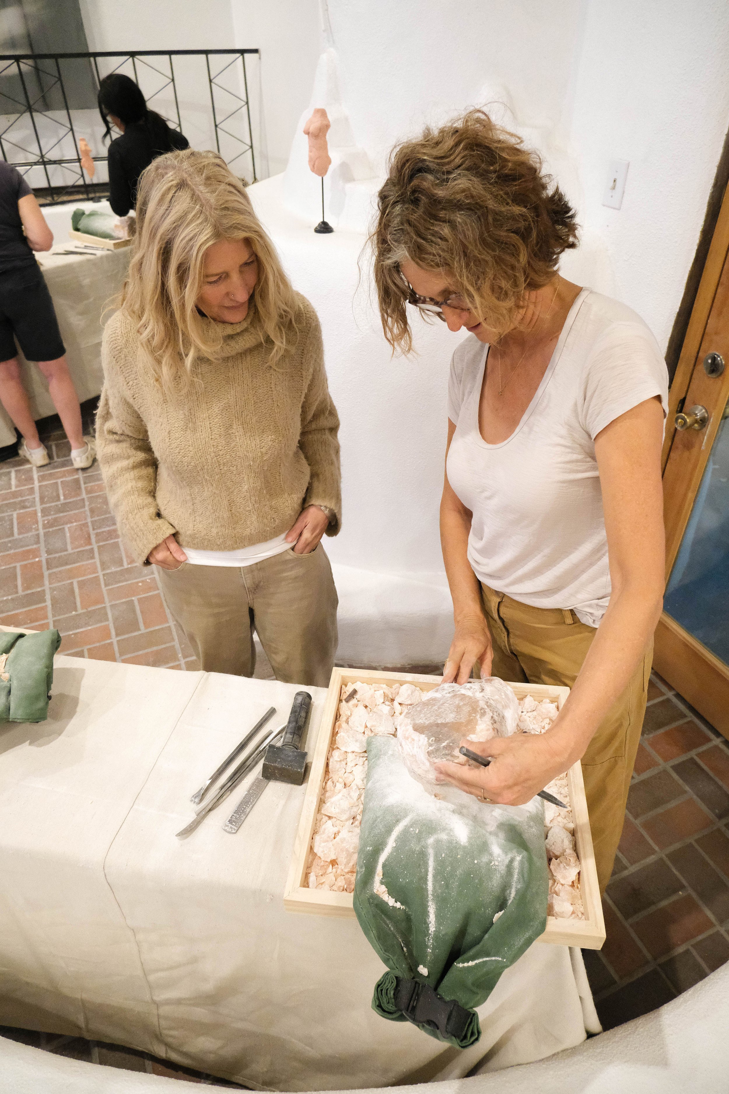 Two women are examining a large fossil embedded in a green rock, surrounded by pink rock fragments, in a museum or exhibit setting.