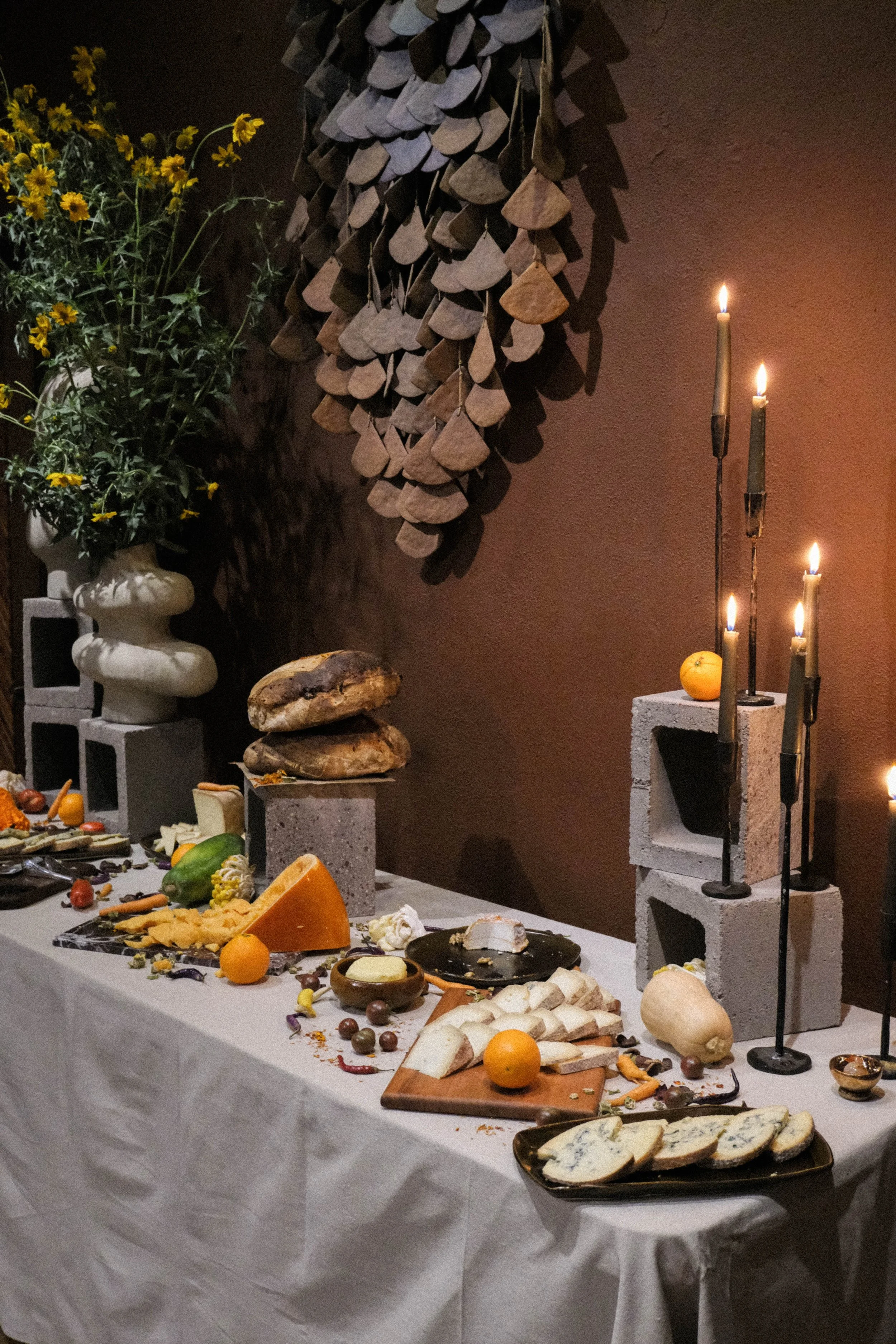 A cheese and bread spread on a white tablecloth with various cheeses, bread, fruits, and chocolates. Candles are lit on candelabras, and decorative objects like cinder blocks and a hanging arrangement of wood slices are in the background, along with 