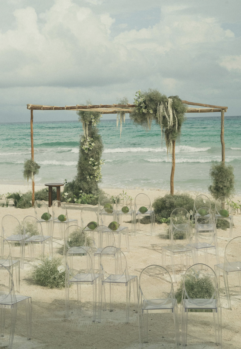 Beach wedding setup with transparent chairs, green floral arrangements, and a bamboo arch decorated with greenery, overlooking the ocean on a cloudy day.