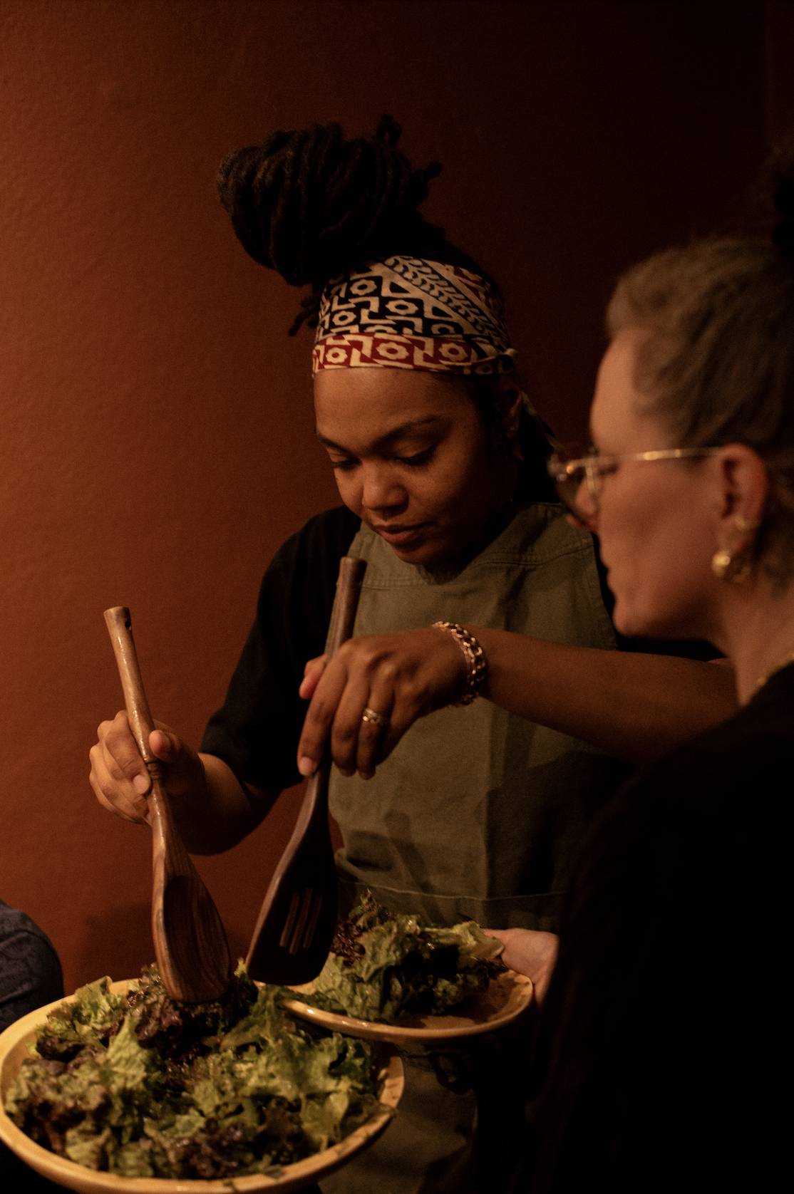 A woman with braids and a colorful headscarf serving salad to a person with glasses at a table.