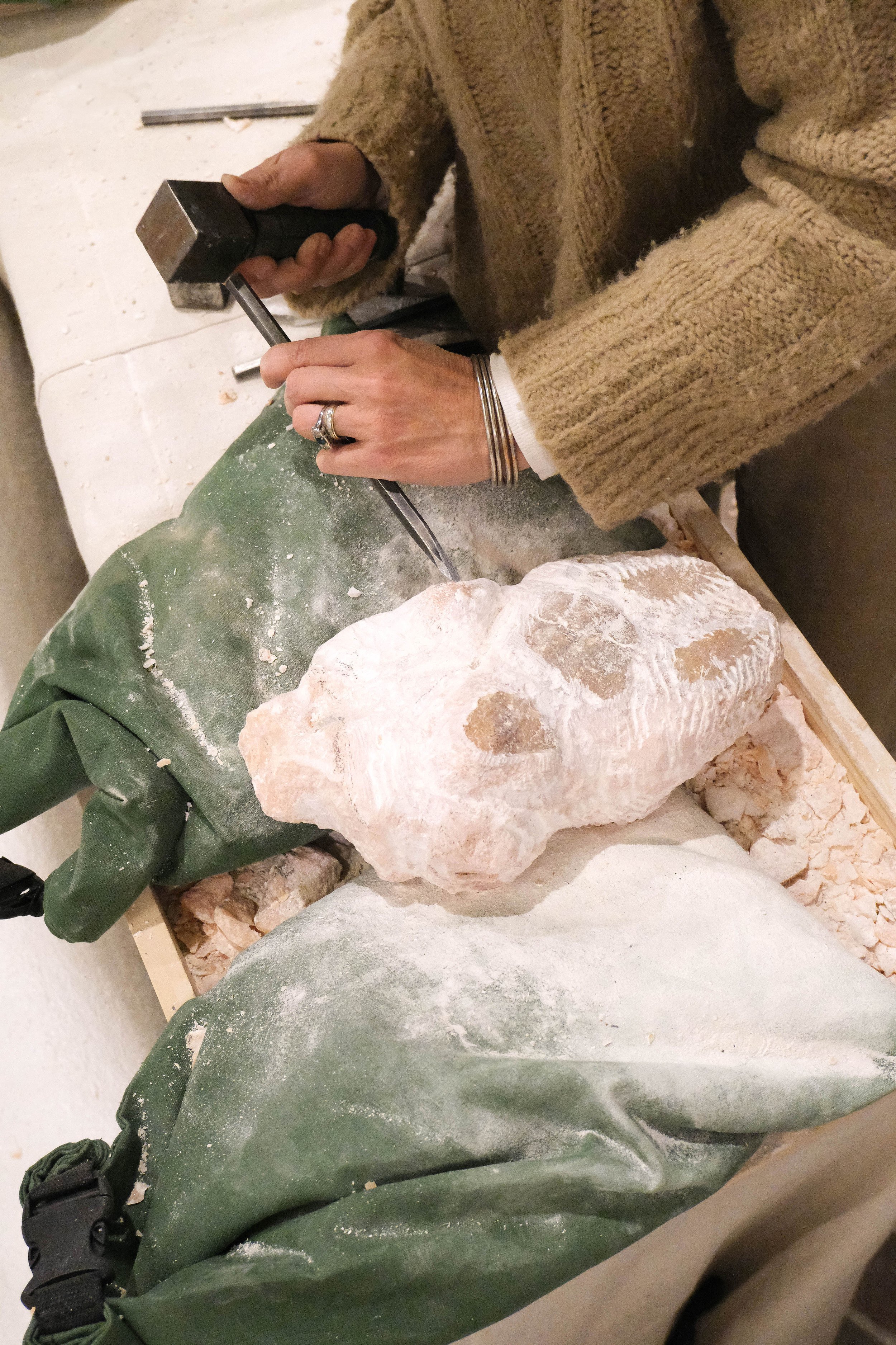 Person sculpting a large stone with a chisel and hammer, wearing a beige sweater and silver rings, working on a green cloth-covered work surface surrounded by stone dust.