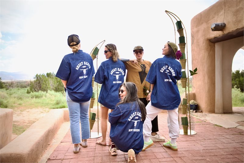 Five women posing outdoors, four wearing matching blue shirts with 'Ghost Ranch' written on the back and a cross logo, and one in the center with a tan jacket and sunglasses, standing in front of an archway with desert landscape in the background.