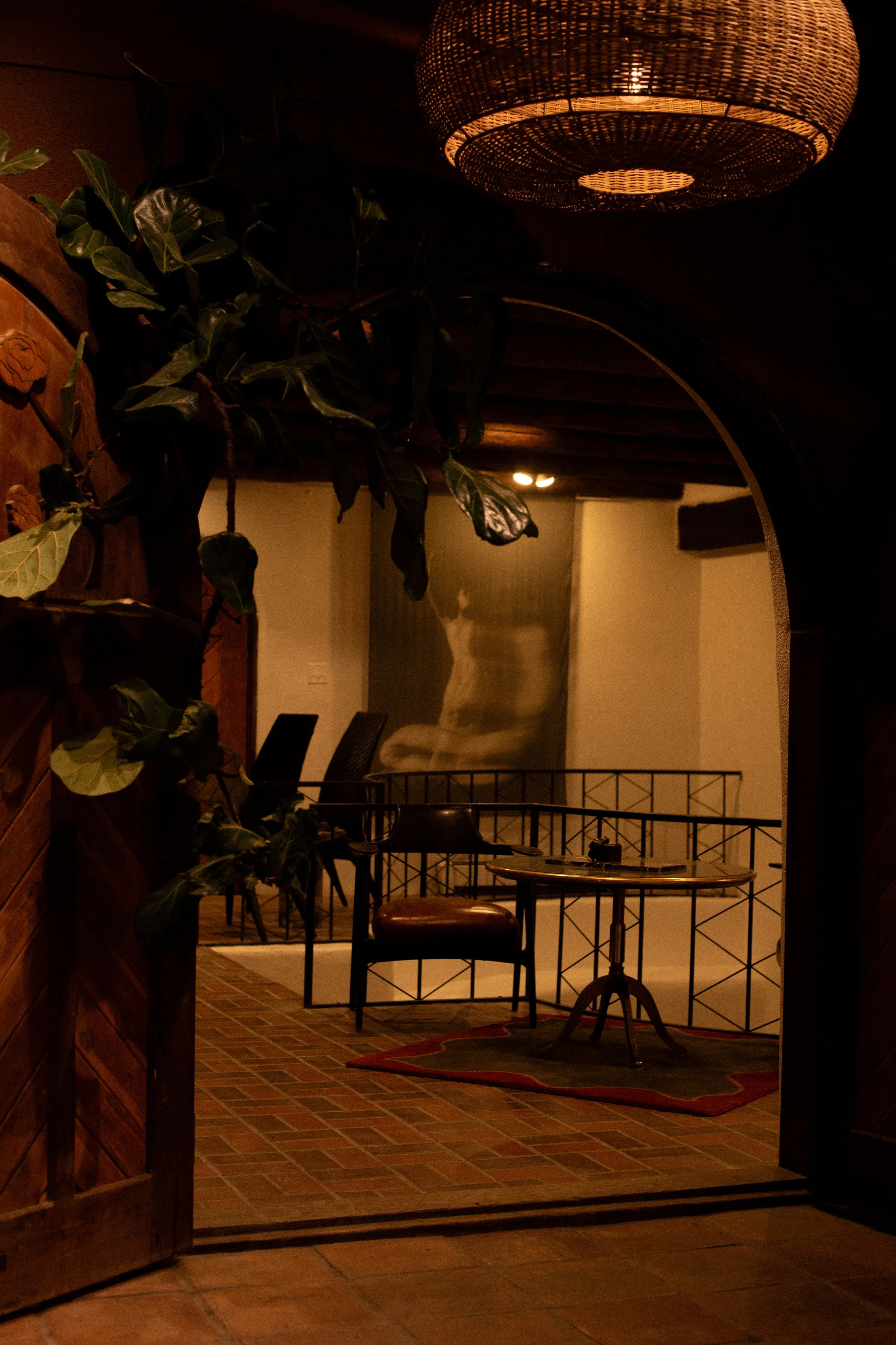 Interior view of a cozy room seen through an arched doorway, featuring a brick floor, a small wooden table with two chairs, a large abstract artwork on the wall, and a ceiling light fixture.